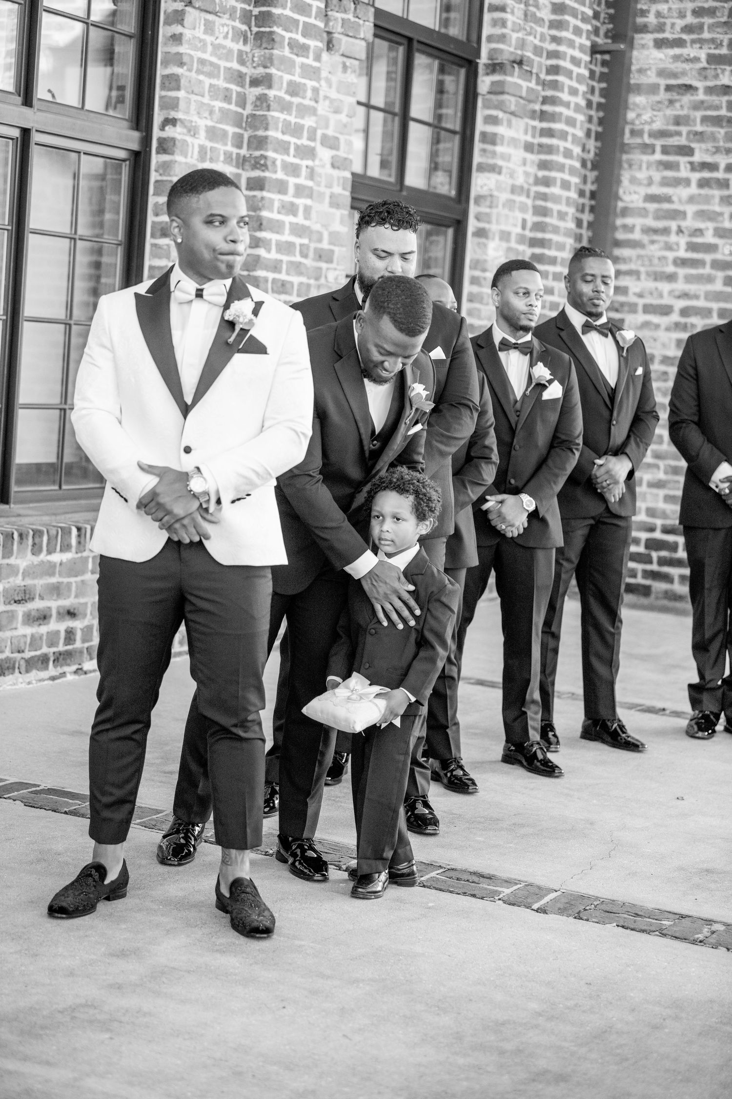 Wedding party members in formal attire lined up against brick building wall.