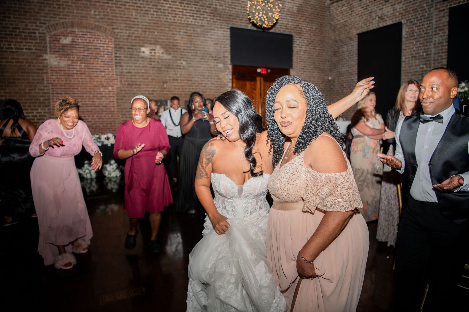 Wedding celebration with guests dancing in formal attire at a brick-walled venue.