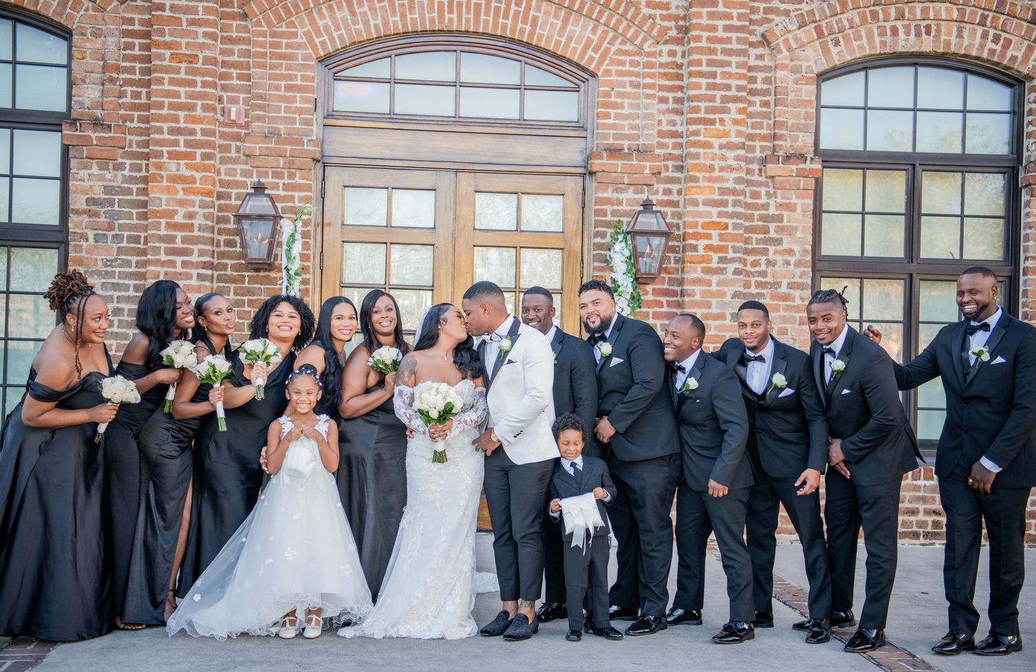 Wide panoramic view of wedding party members seated and standing against industrial brick backdrop.