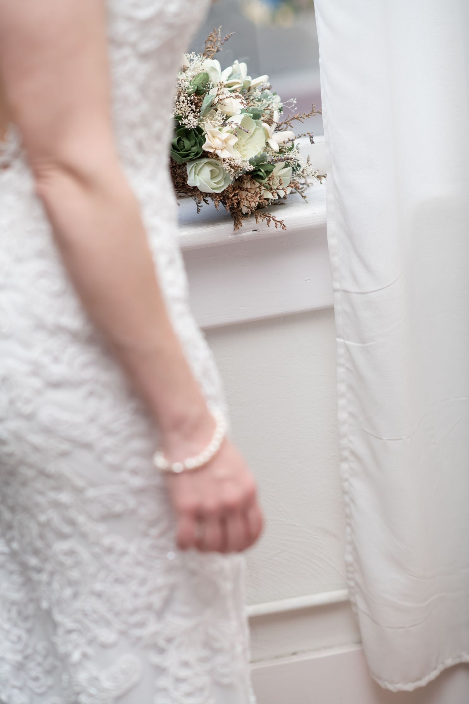 Close up of a delicate white and green bridal bouquet next to a lace wedding dress.