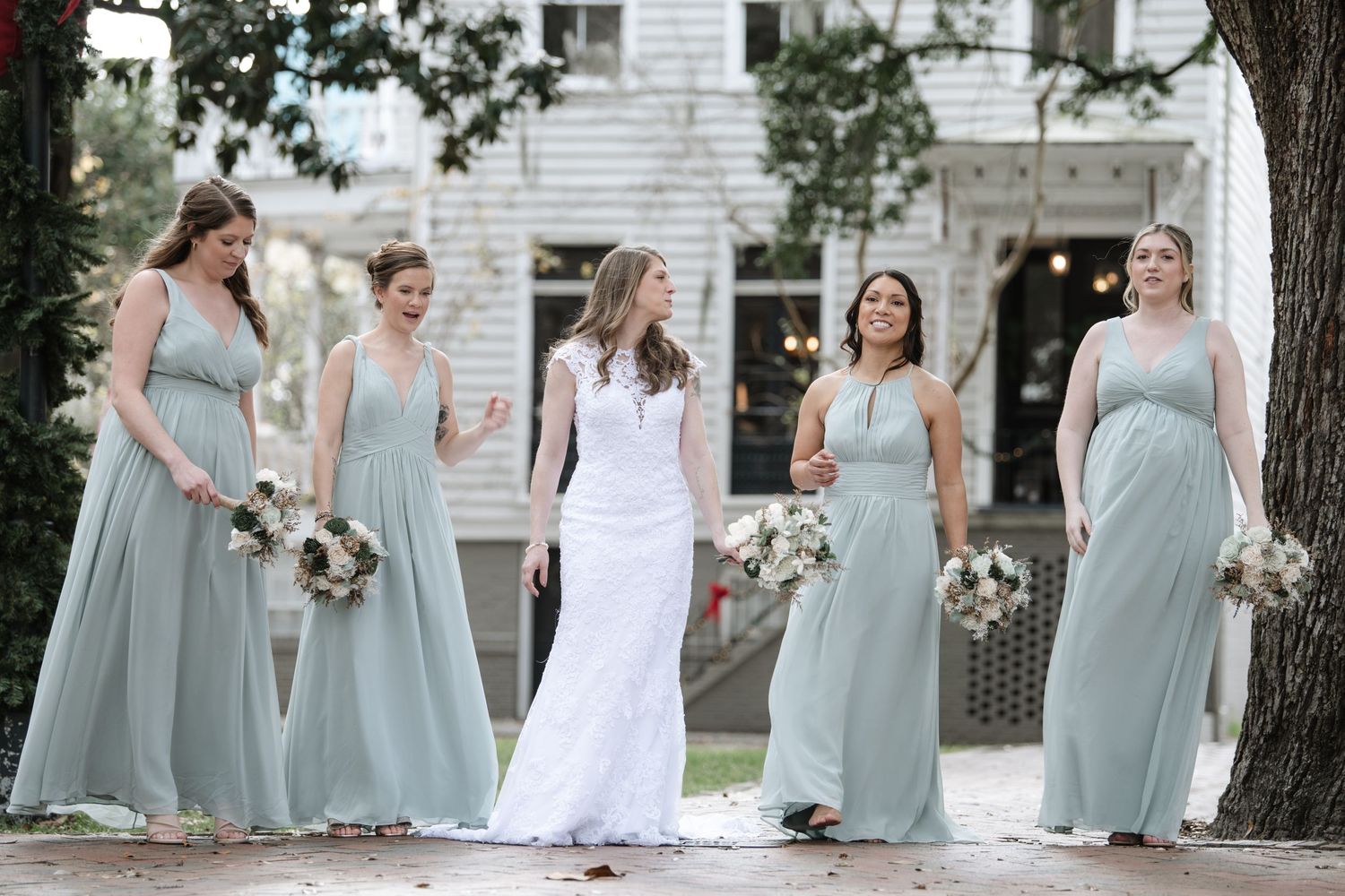 Bridesmaids in sage green dresses pose with bouquets on a white house porch.