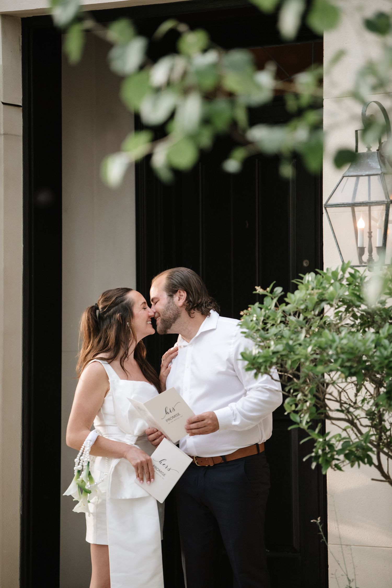 A romantic couple shares a tender kiss on a porch with white columns and lush greenery in the background.