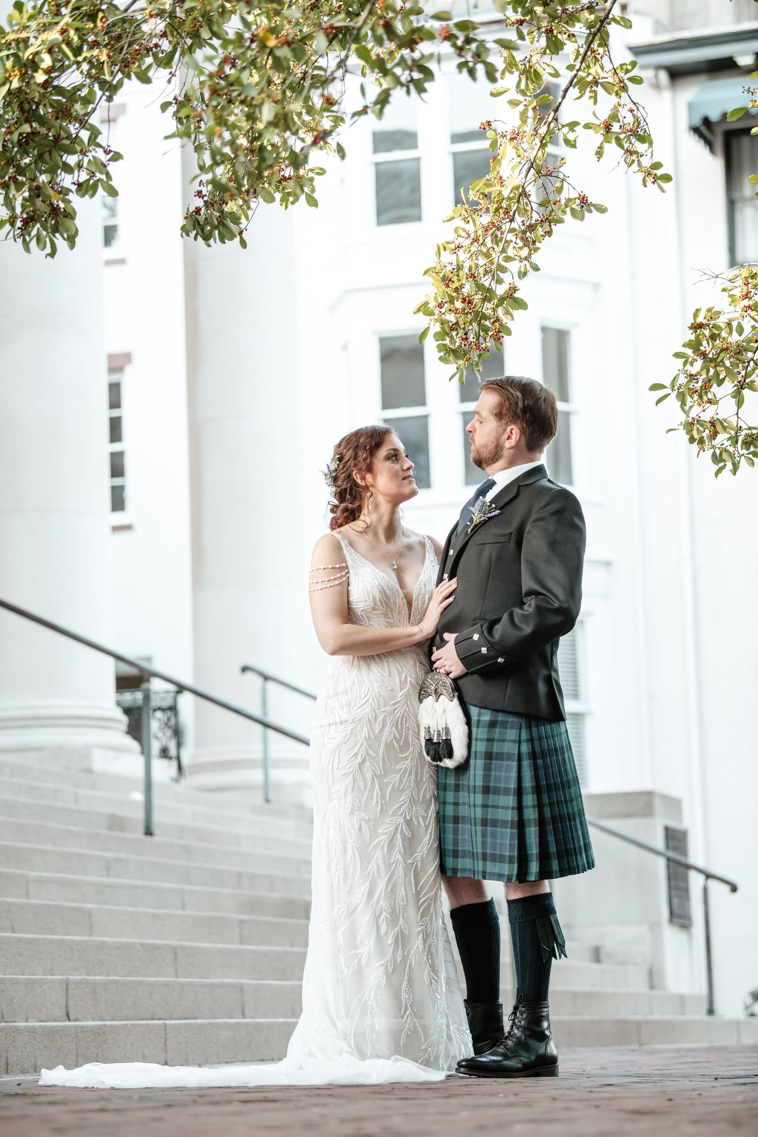 Couple in wedding attire posing on white steps under autumn leaves.