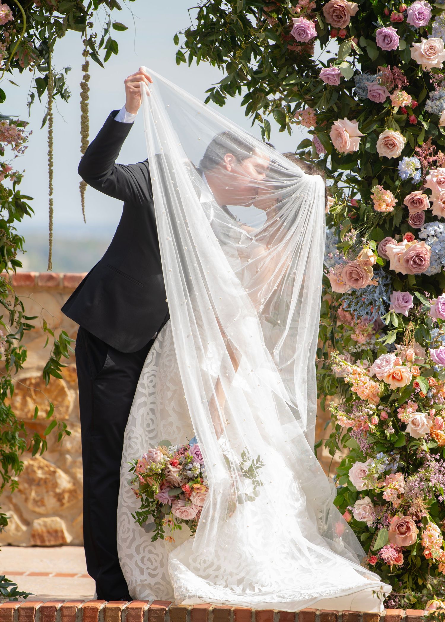 Wedding couple posing with flowing white veil against a backdrop of climbing pink roses.