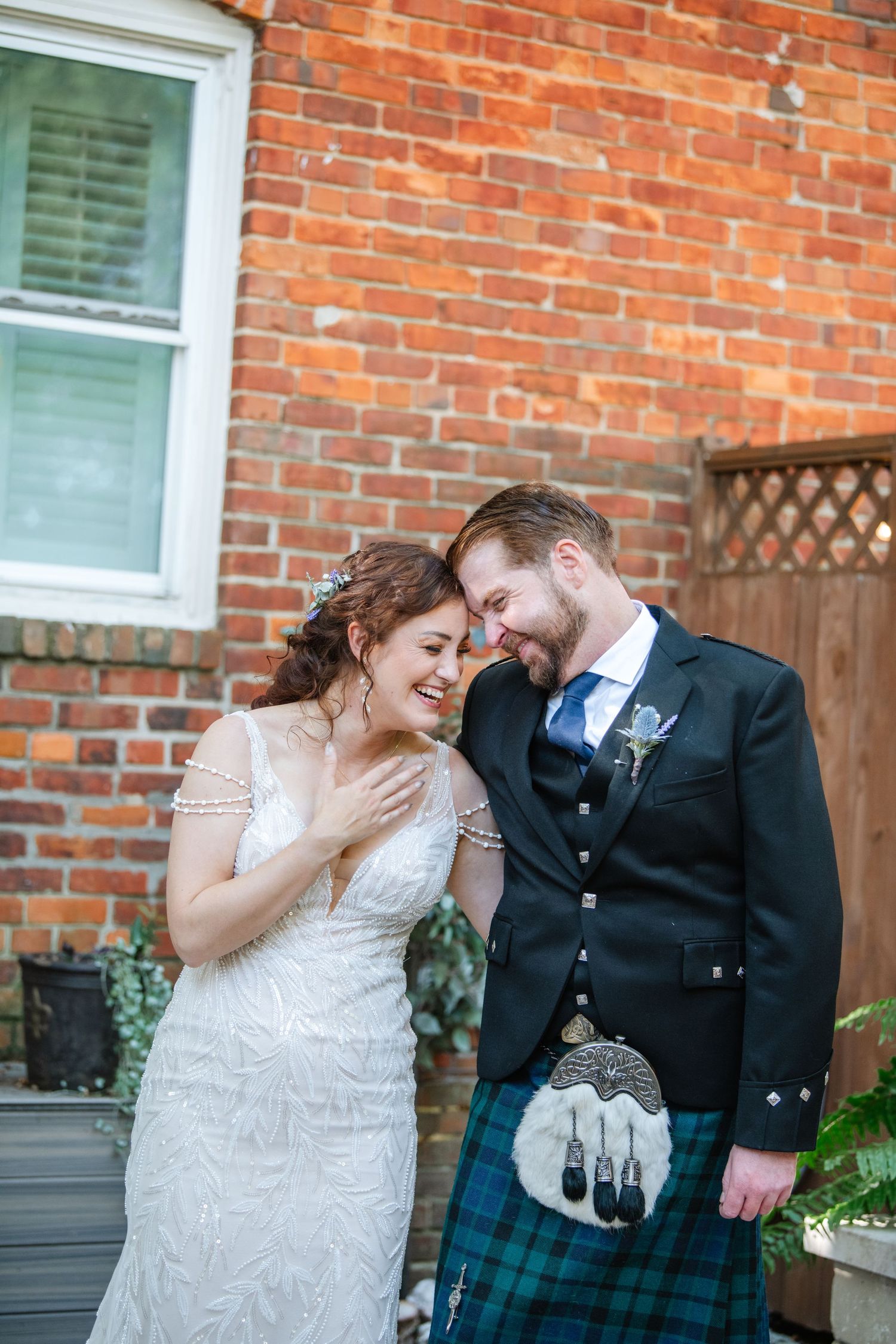 Candid moment of couple sharing laughter against red brick wall with groom in traditional Scottish attire.
