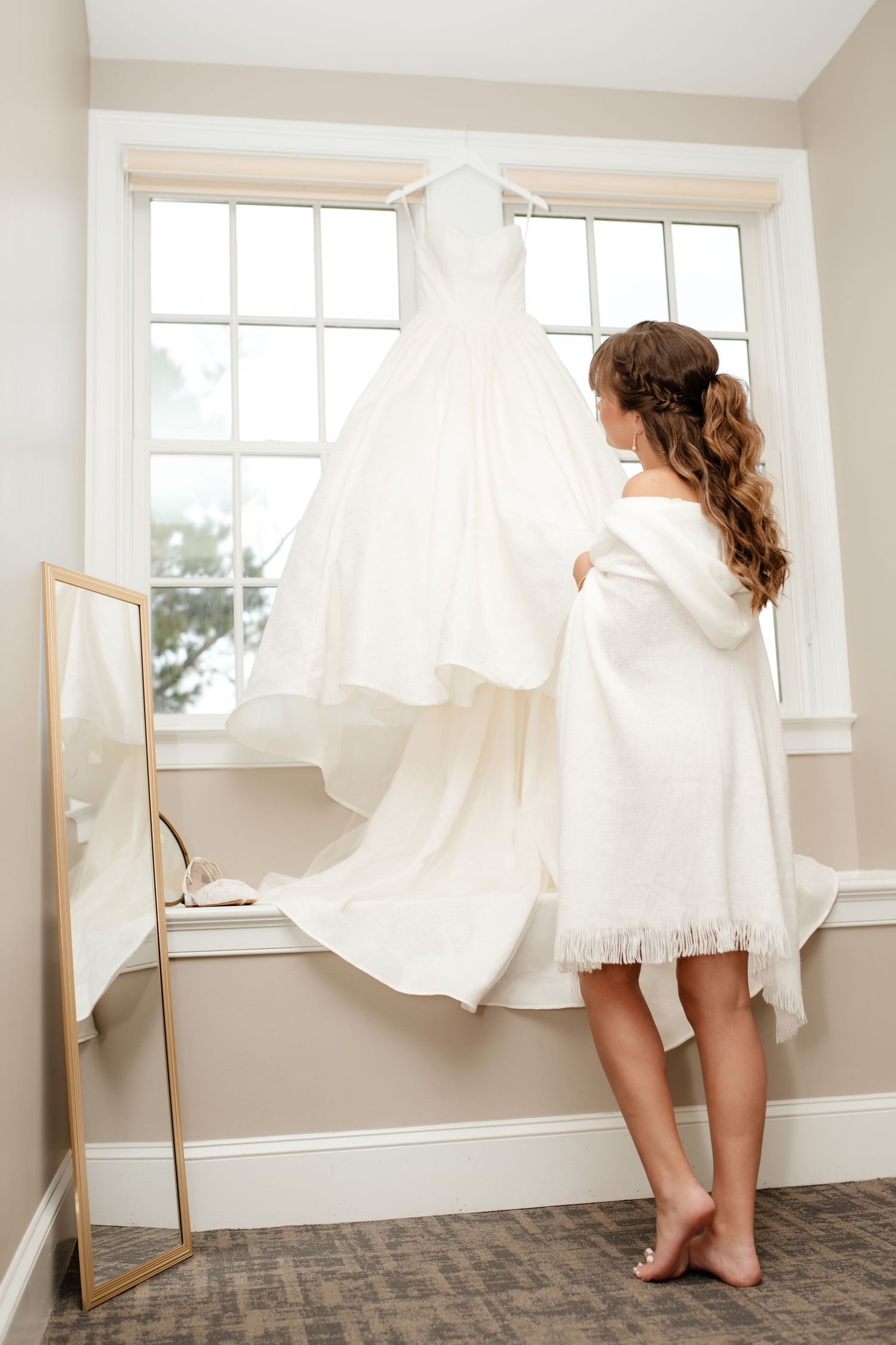 White wedding dress hanging by window with bridal preparation scene.