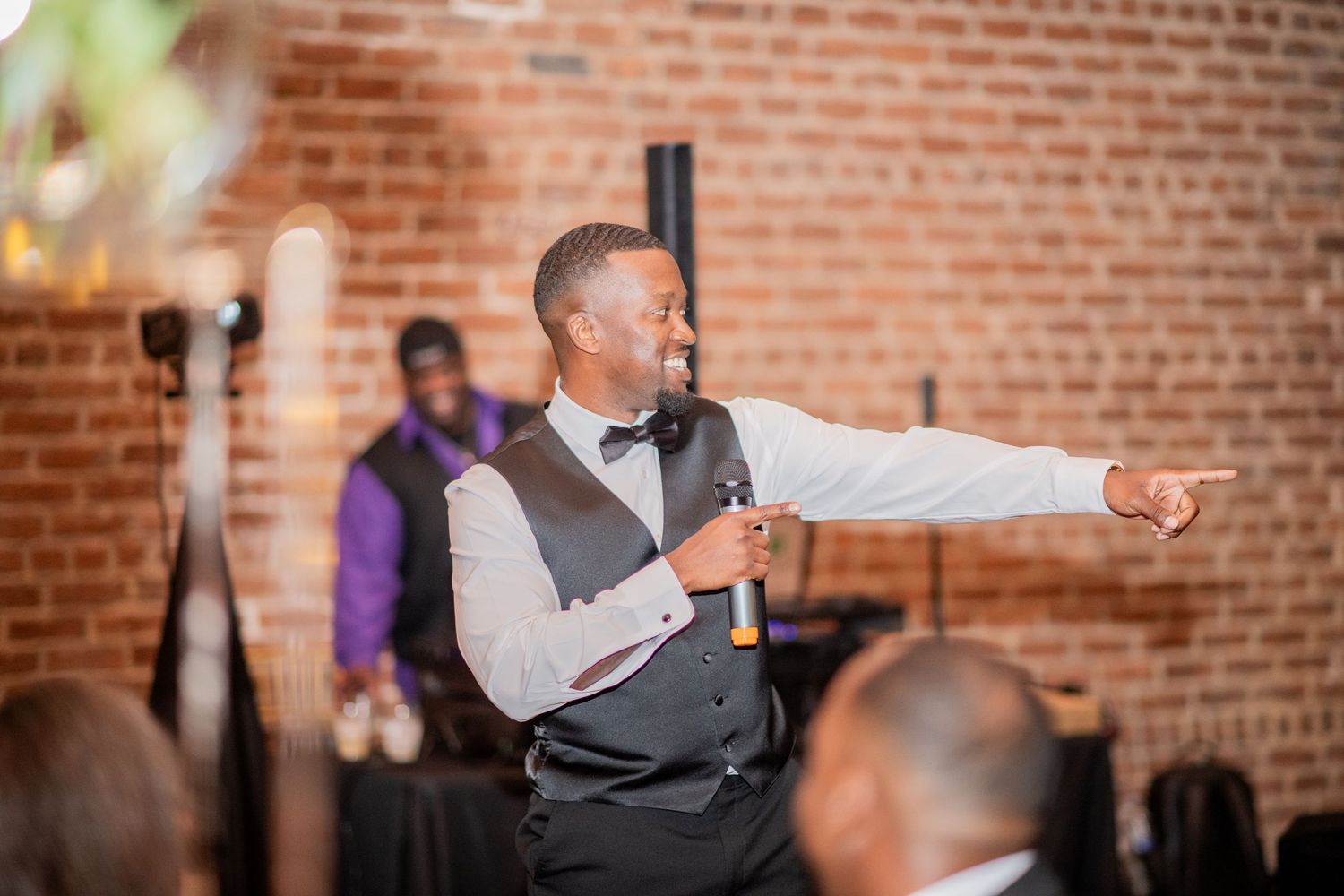 A speaker in formal attire gestures during a speech at an indoor venue with exposed brick walls.
