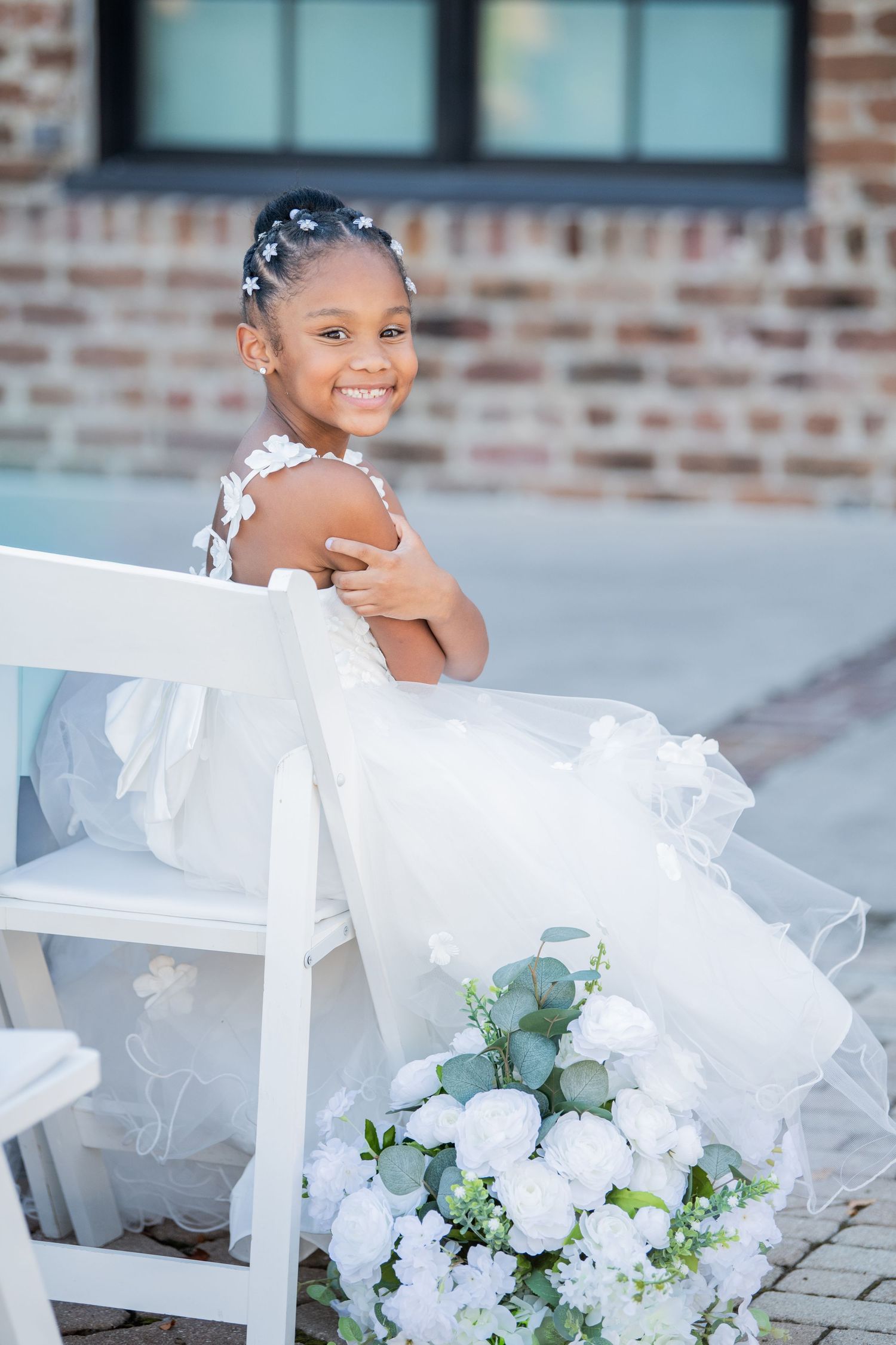 Young attendant in white dress sitting on chair next to floral arrangement.