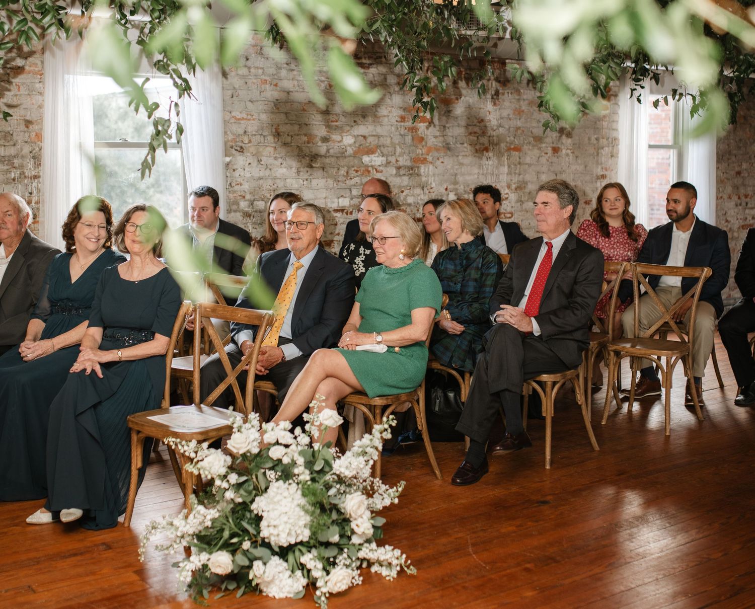 Wedding ceremony guests seated in wooden chairs at a rustic brick venue with floral decorations.