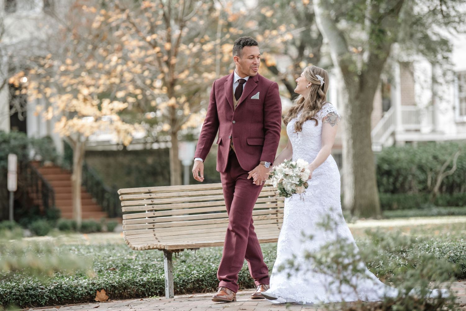 A couple shares romantic moments in a garden setting wearing burgundy suit and white lace gown.