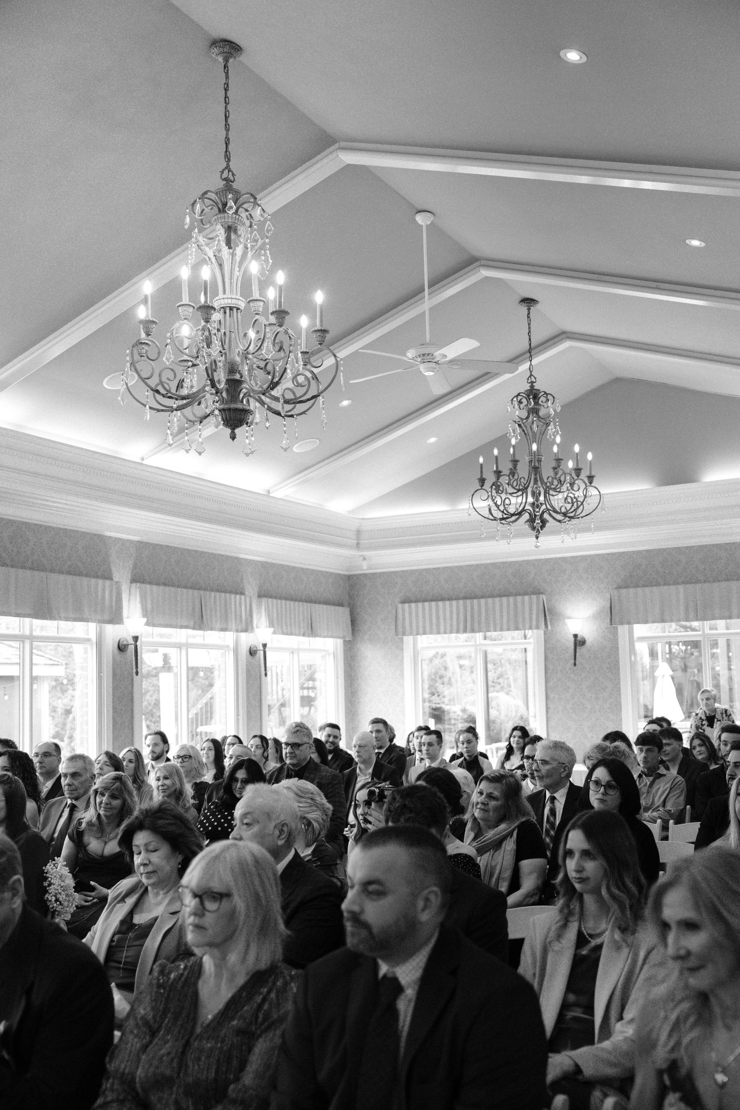 Black and white photo of wedding guests seated in an elegant venue with chandeliers during a ceremony.