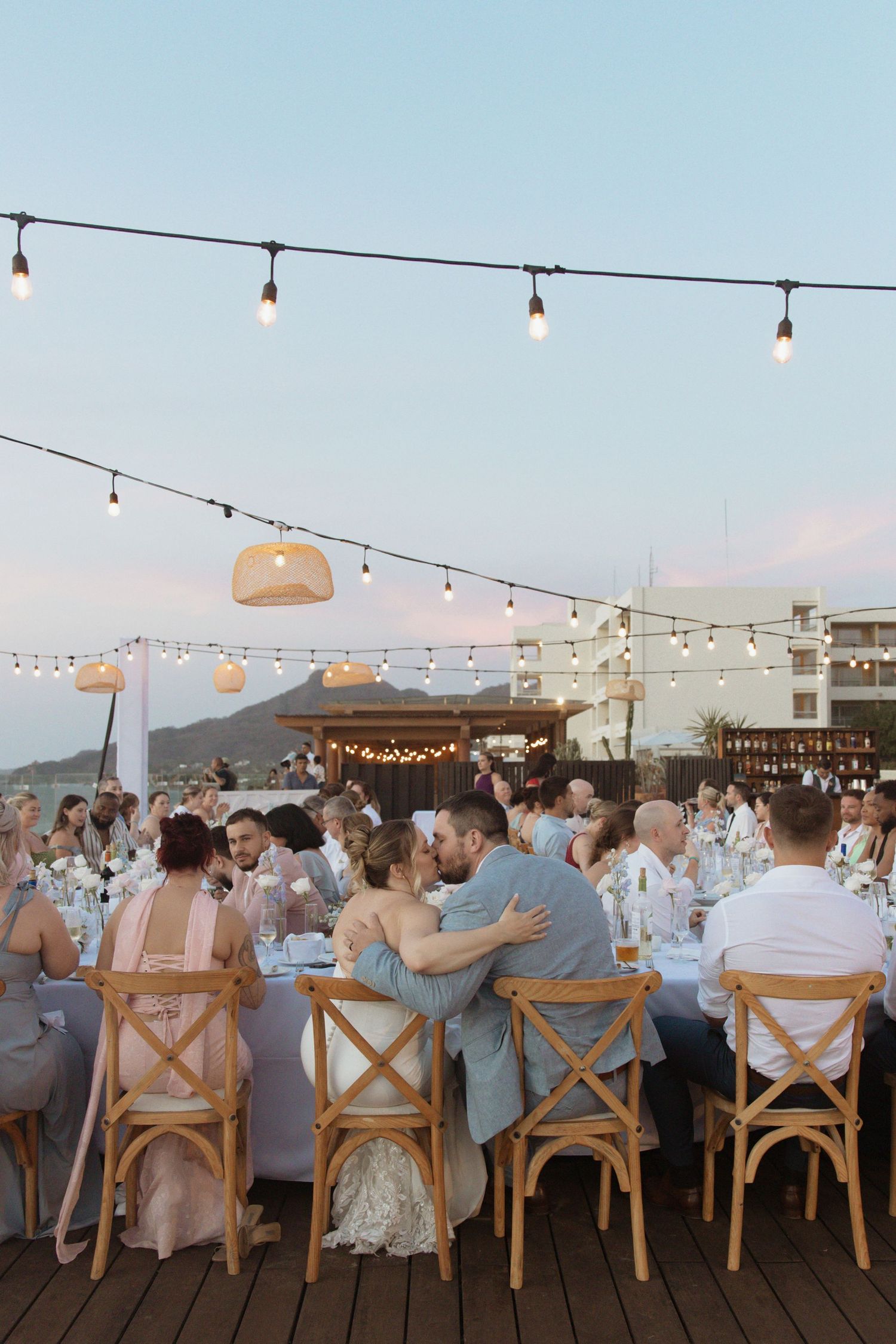 Wedding reception dinner on outdoor terrace with string lights and guests at decorated tables.