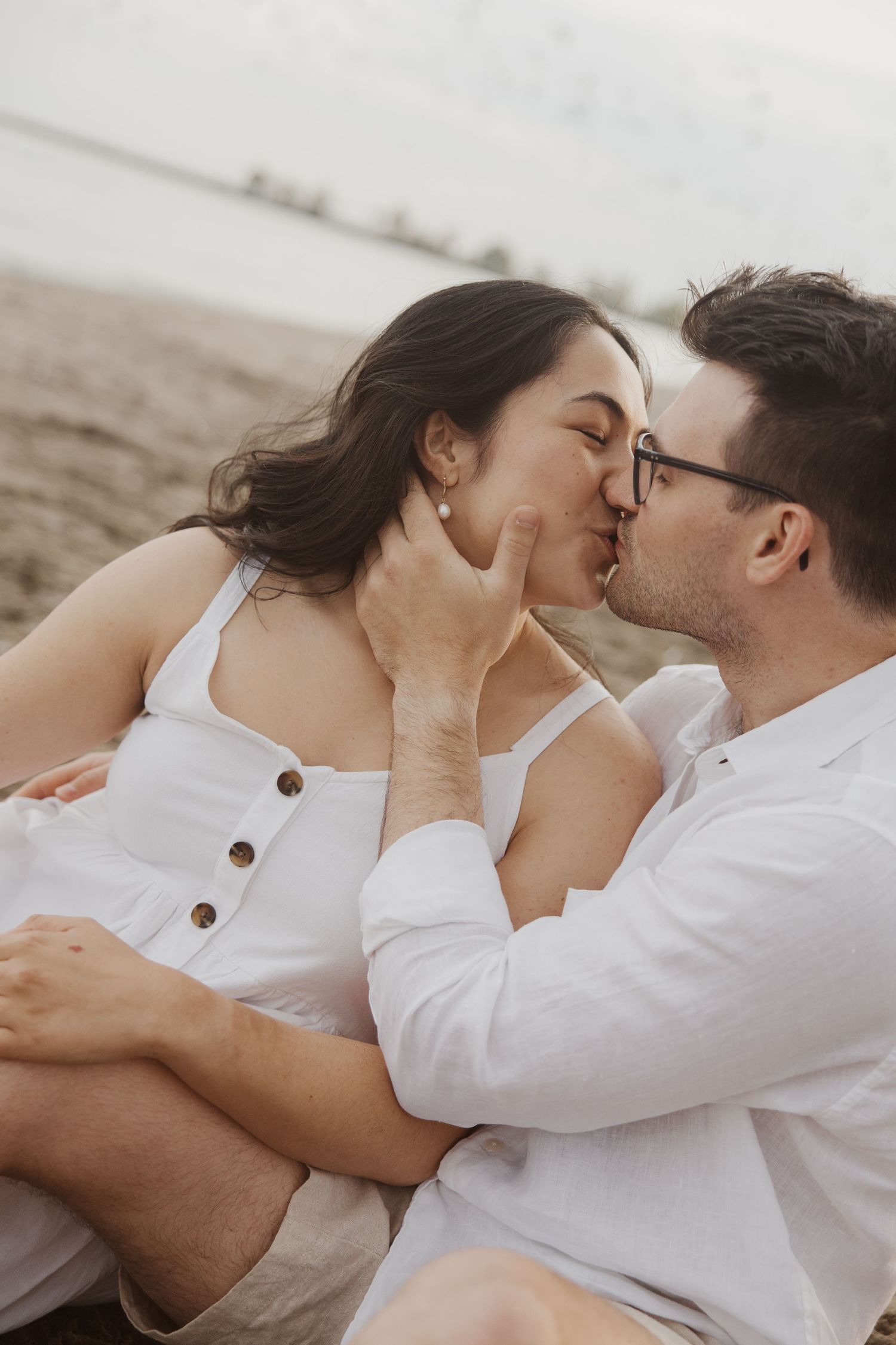 Couple sharing a kiss while sitting on sandy beach near water.