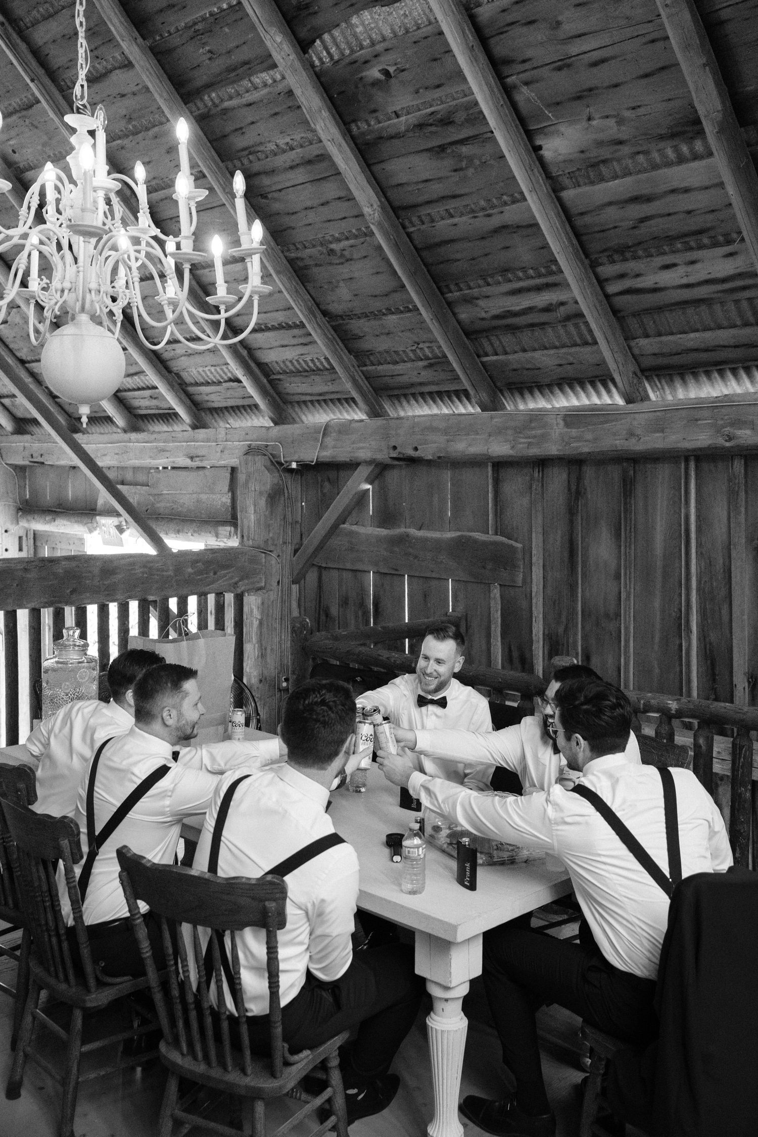 Black and white photo of groomsmen toasting at a table inside a rustic barn venue.