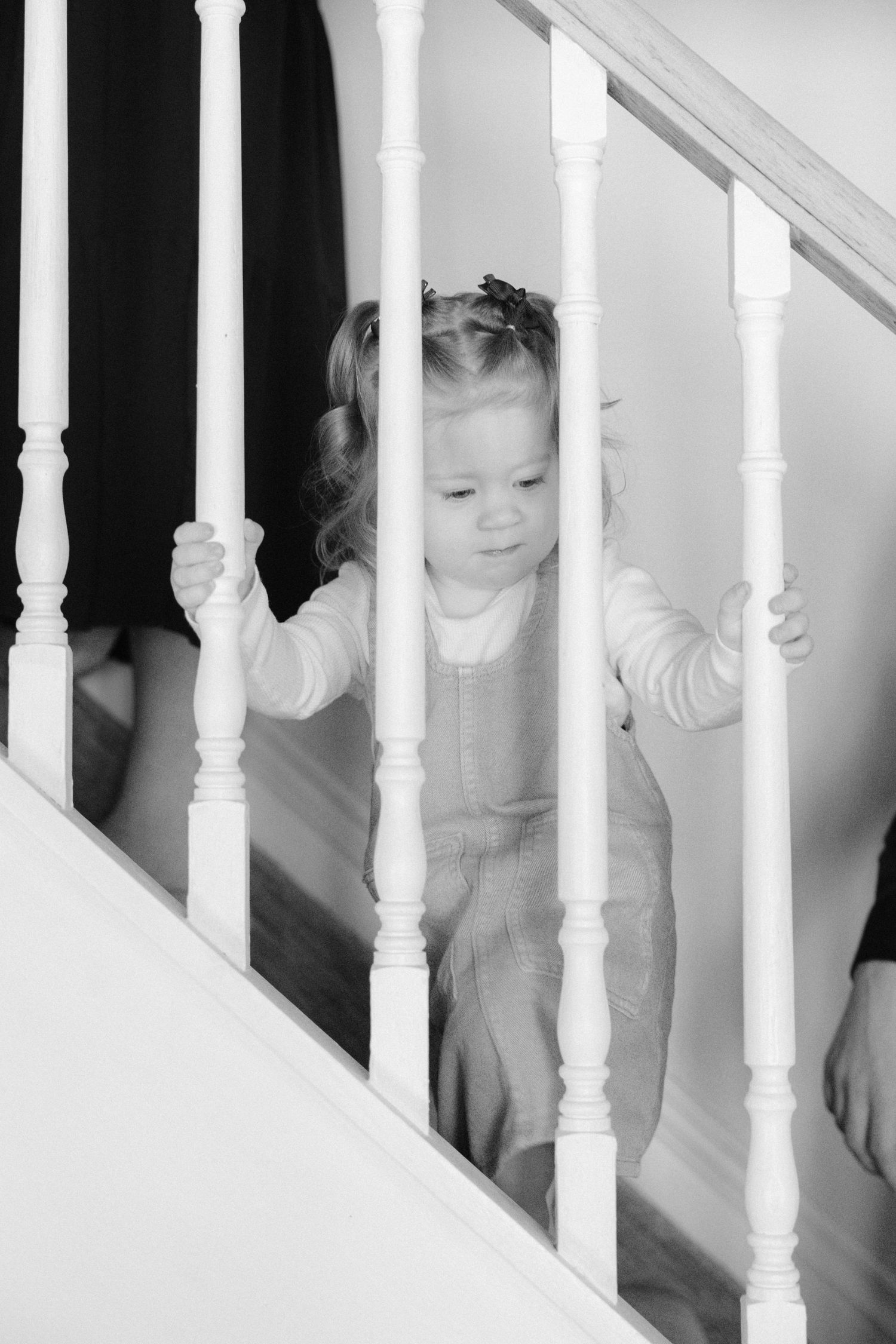 Black and white photo of a young toddler girl gripping white stair railings while looking down.