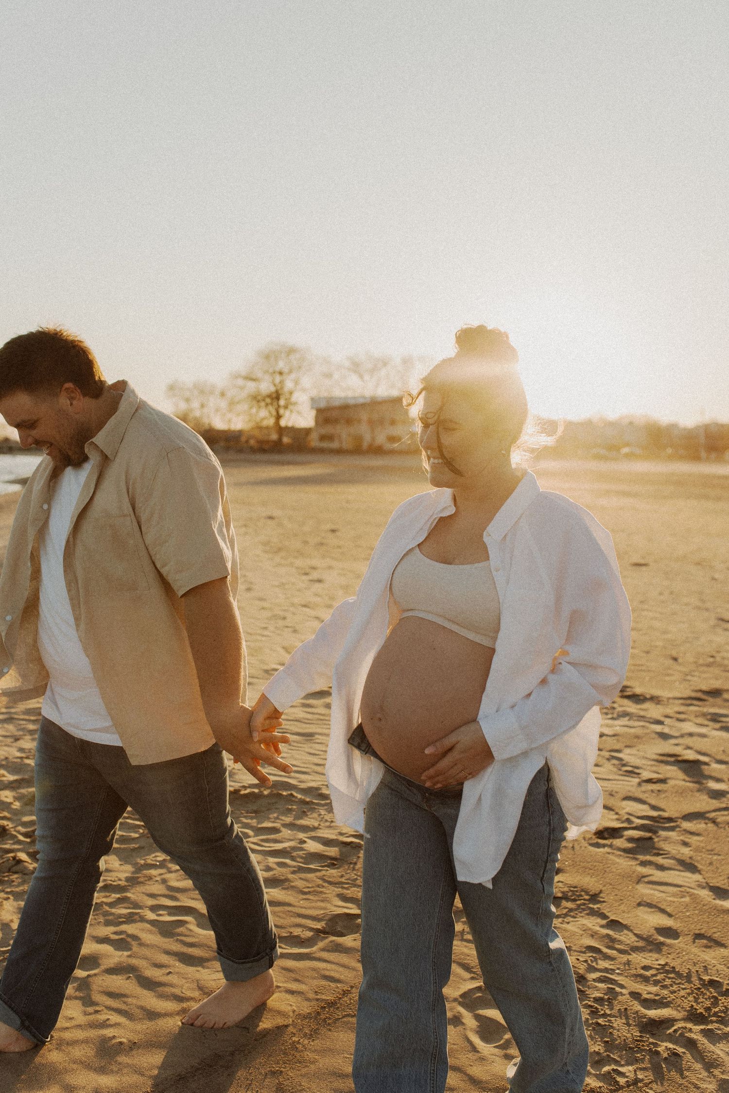 Expecting couple walking hand in hand on a sandy beach during a warm golden sunset.