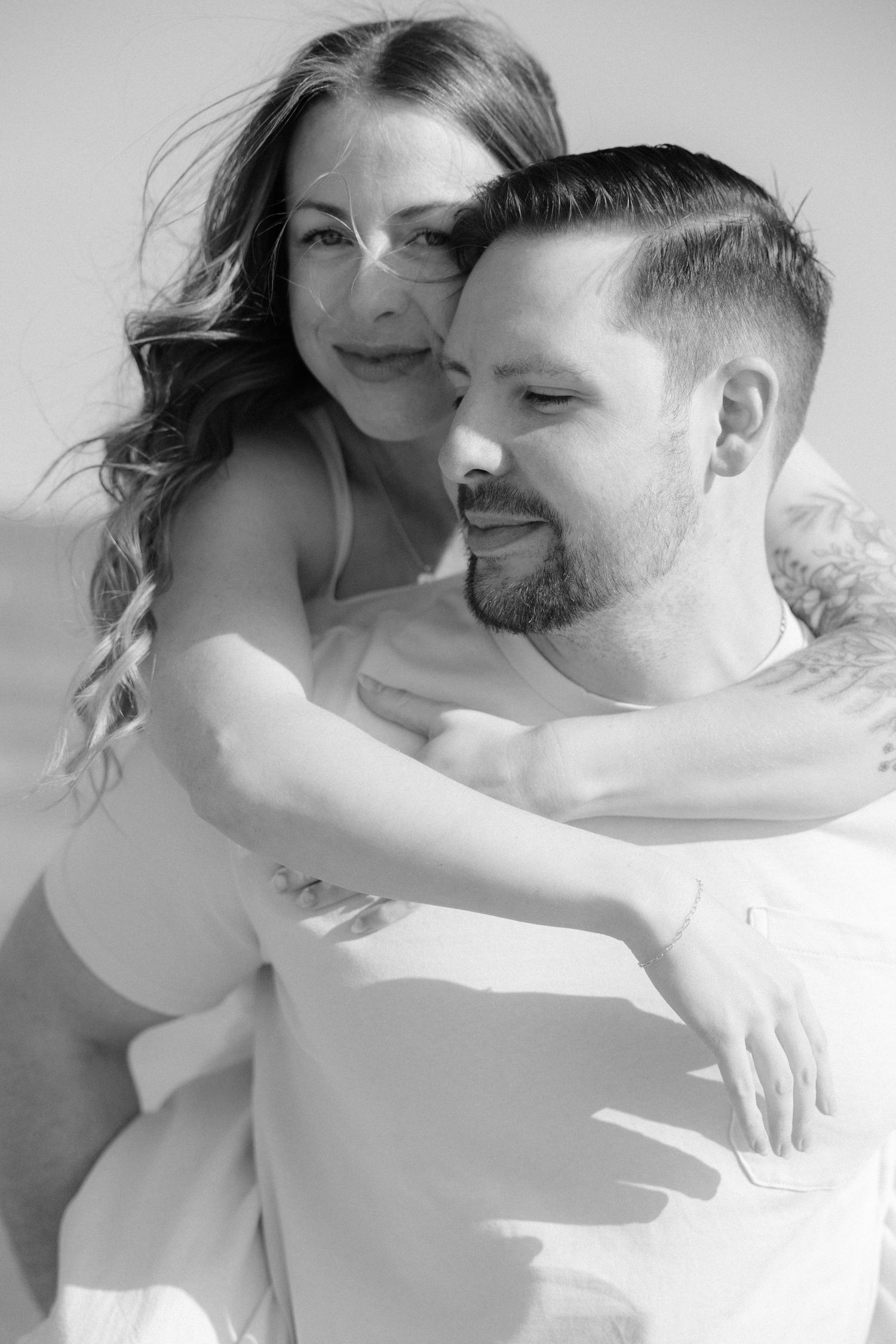 Black and white romantic portrait of woman embracing man from behind on sunny day.