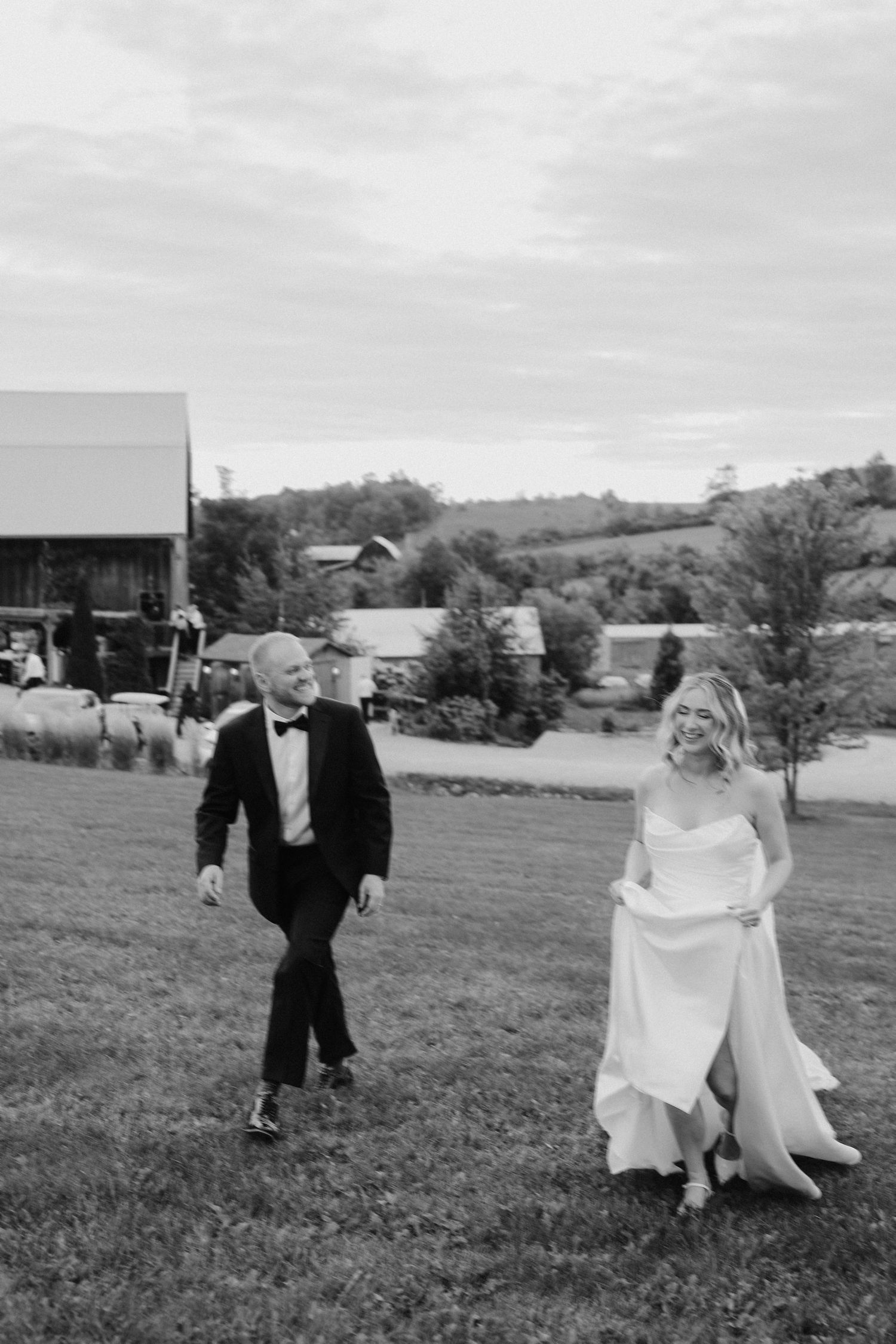 Black and white photo of bride and groom walking and laughing across green field with rustic barn backdrop.