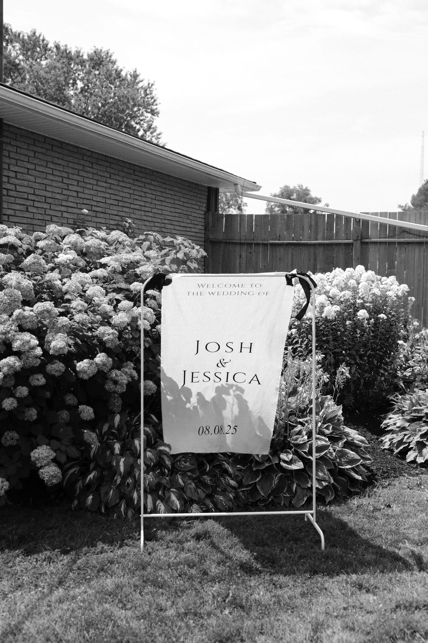 Black and white welcome sign for Josh and Jessica wedding dated 08.06.25 displayed among hydrangea bushes.