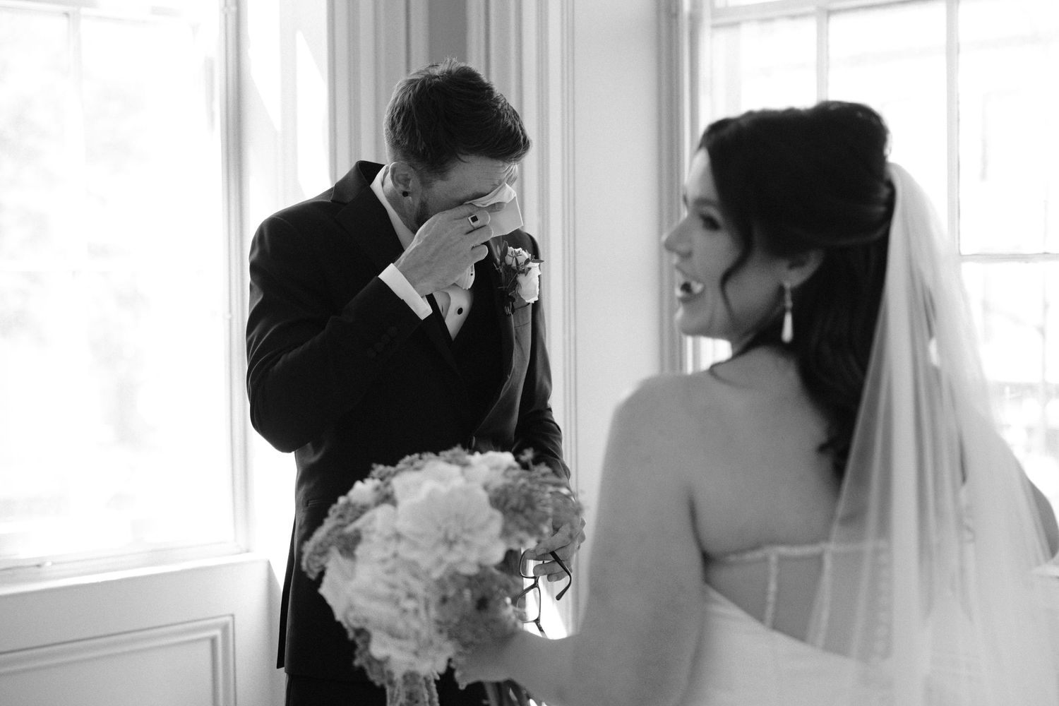 Emotional groom covering his face while bride smiles during their first look at wedding ceremony.