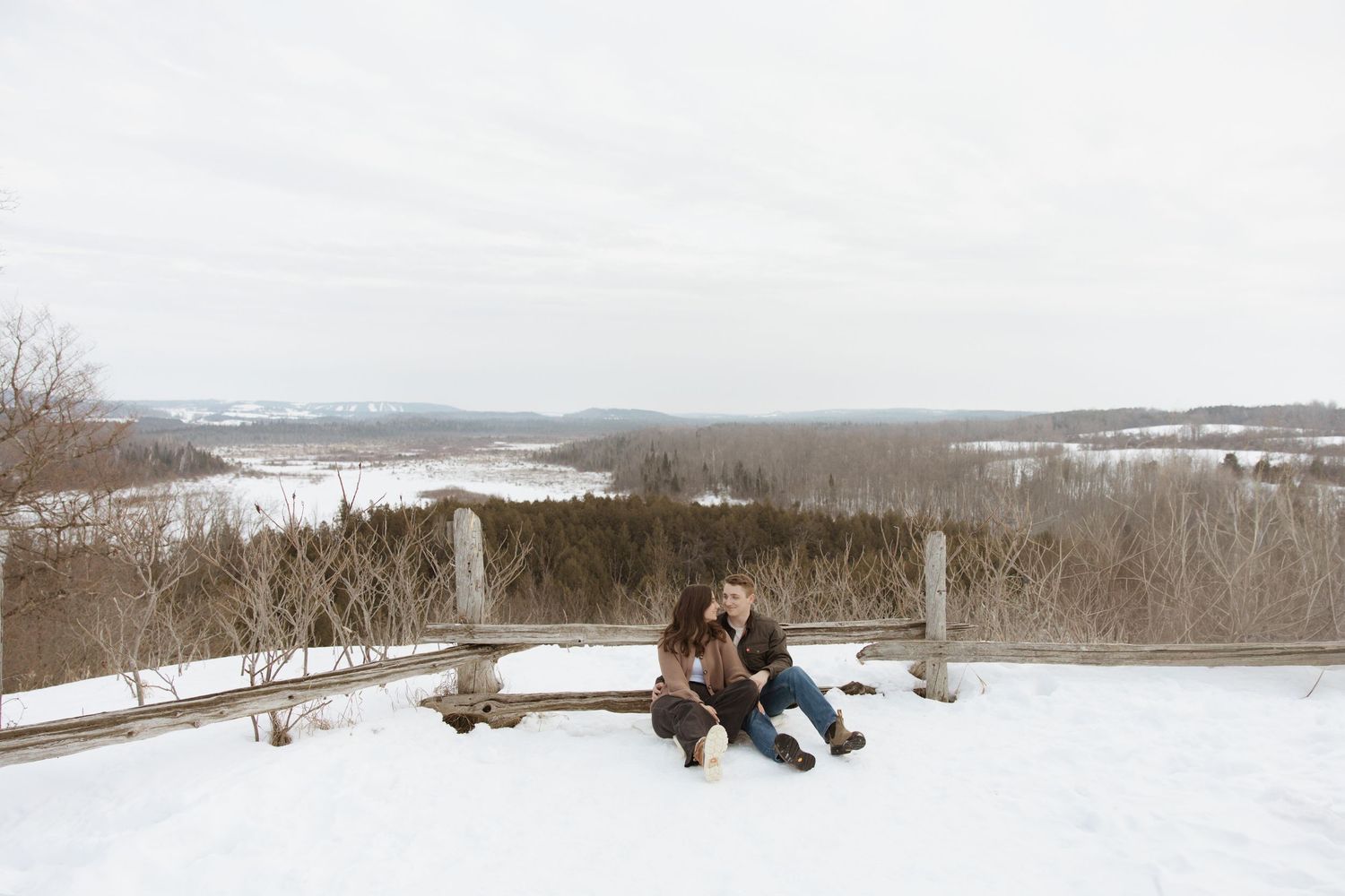 A couple sits together on a snowy bench overlooking a vast winter landscape with bare trees.