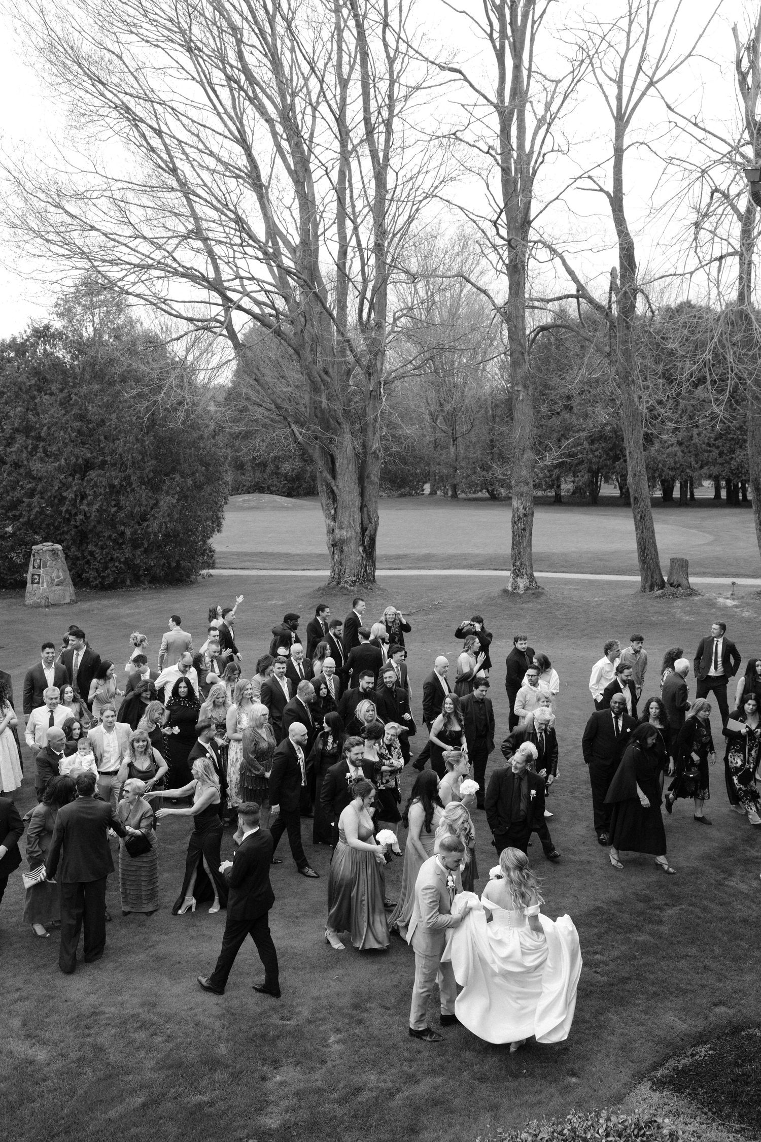 Aerial black and white photo of wedding guests mingling outdoors on a lawn surrounded by bare trees.