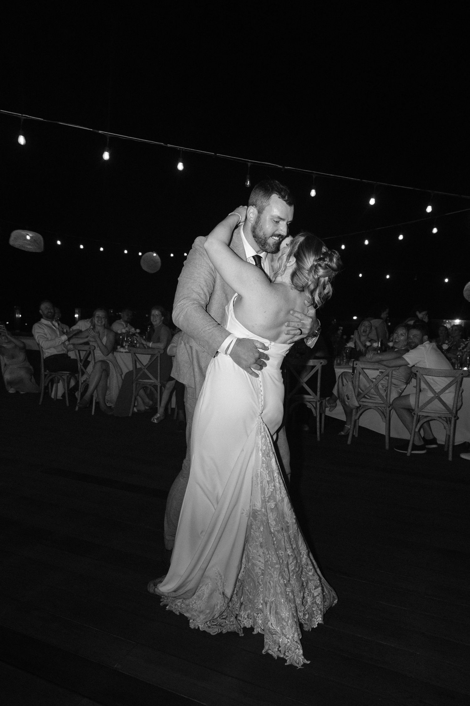 Black and white photo of bride and groom sharing romantic first dance under string lights at outdoor wedding reception.