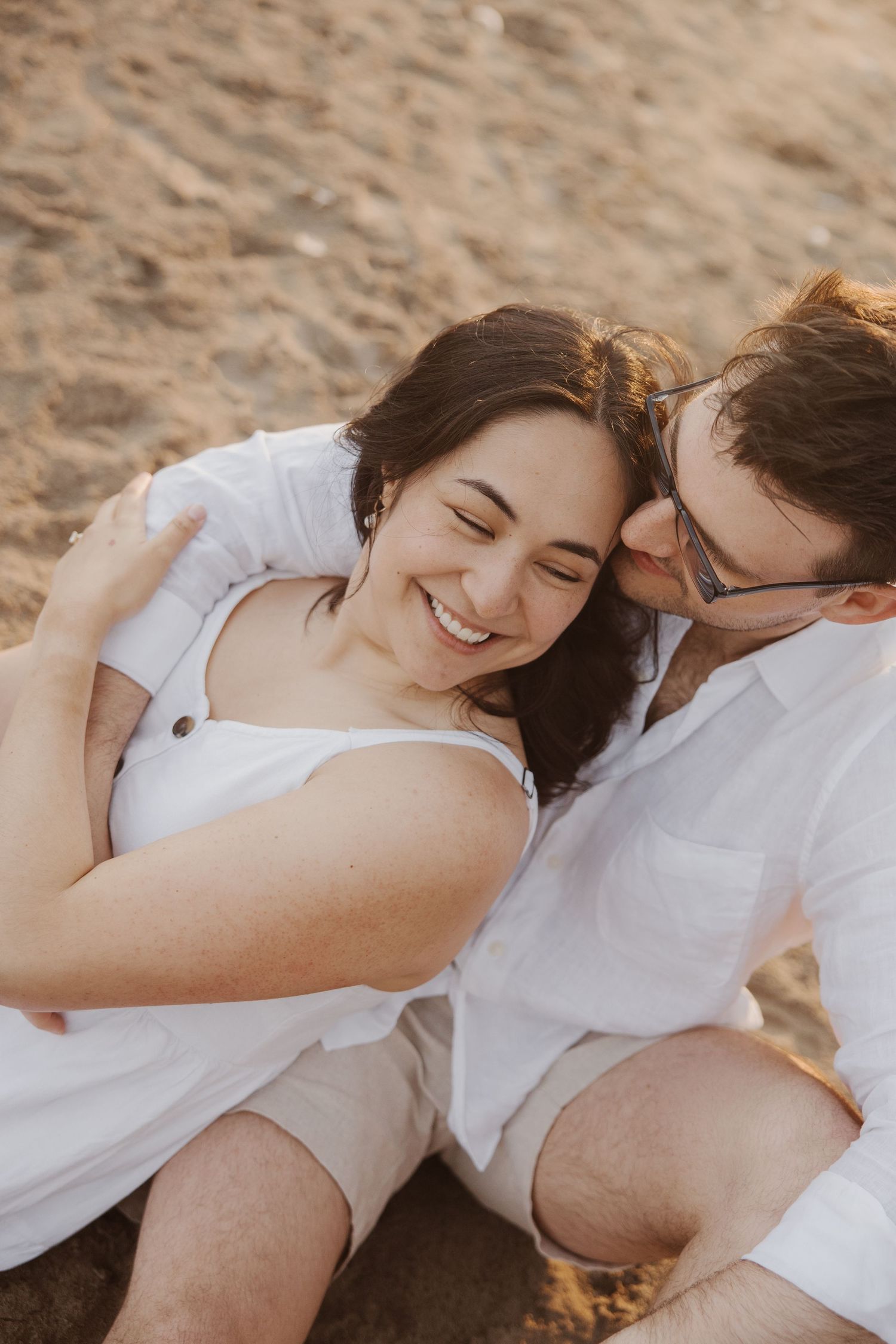 Happy couple laughing and cuddling together while sitting on sandy beach.