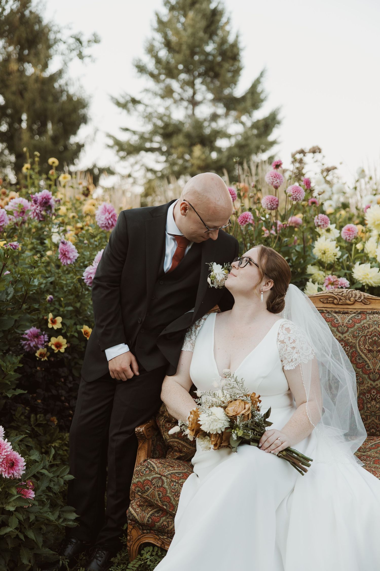 Groom leaning toward seated bride holding bouquet surrounded by colorful dahlia flowers in garden wedding setting.