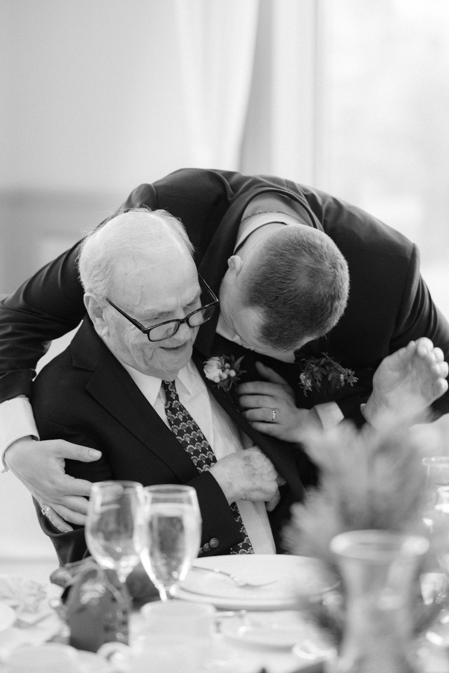 Emotional black and white photo of groom embracing an elderly grandfather at the wedding reception table.