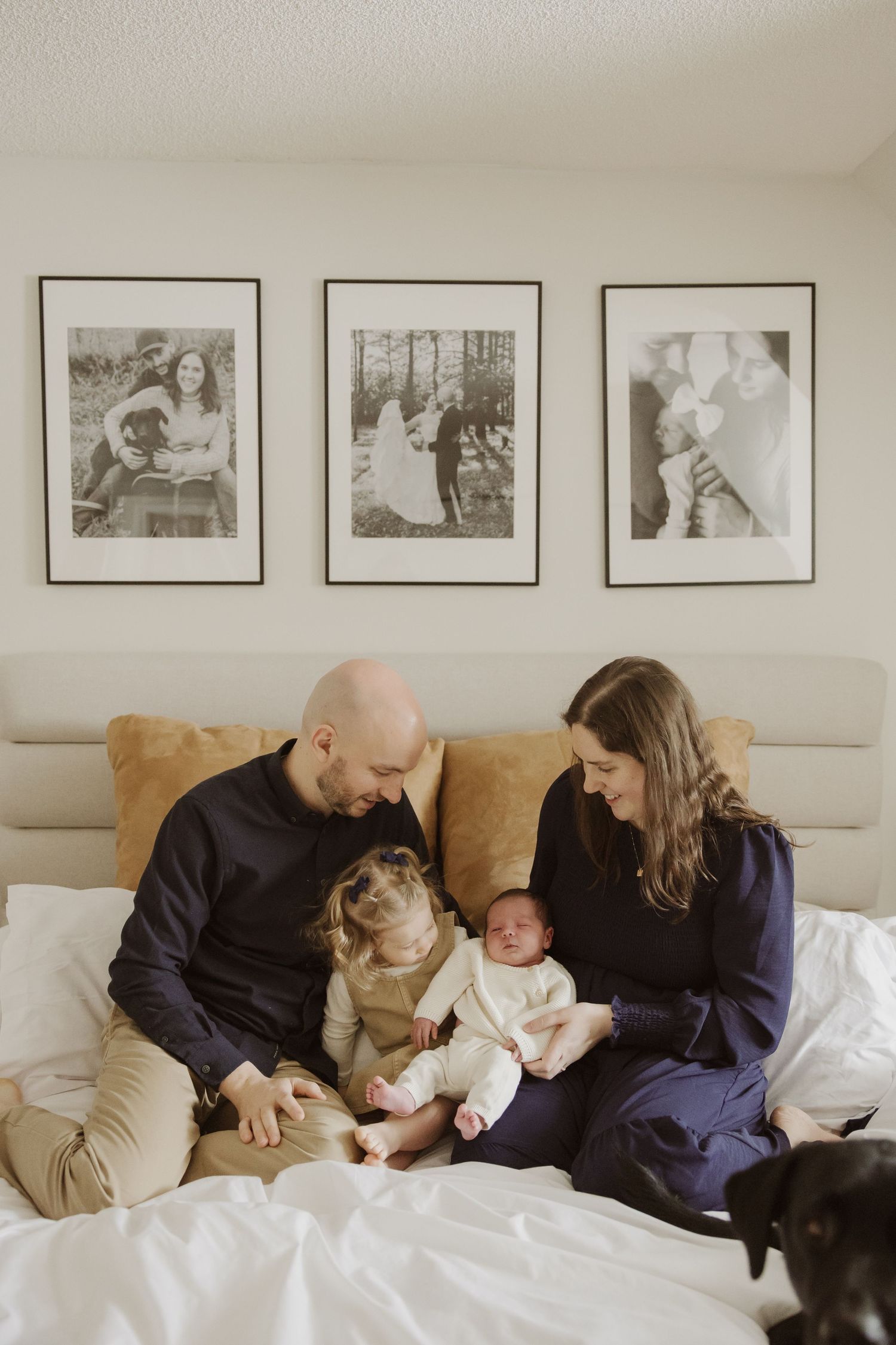 Family of four with a toddler, newborn, and black dog sitting together on a white bed at home.