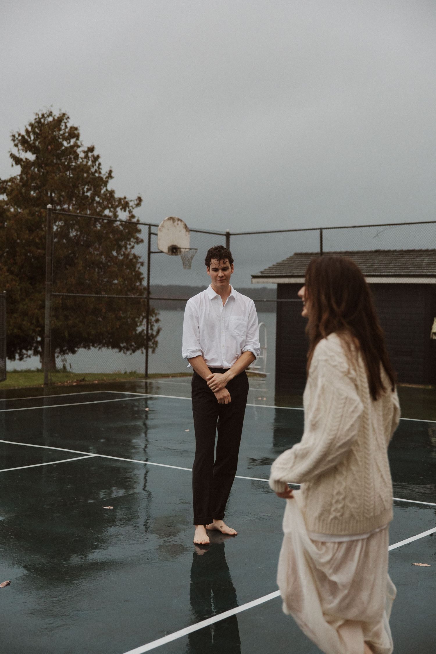 Couple on wet basketball court in rain, woman in lace dress holding racket.