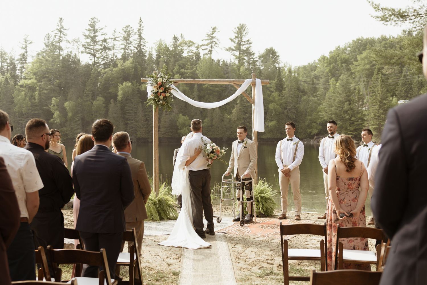 Outdoor lakeside wedding ceremony with wooden arch and guests seated on chairs.