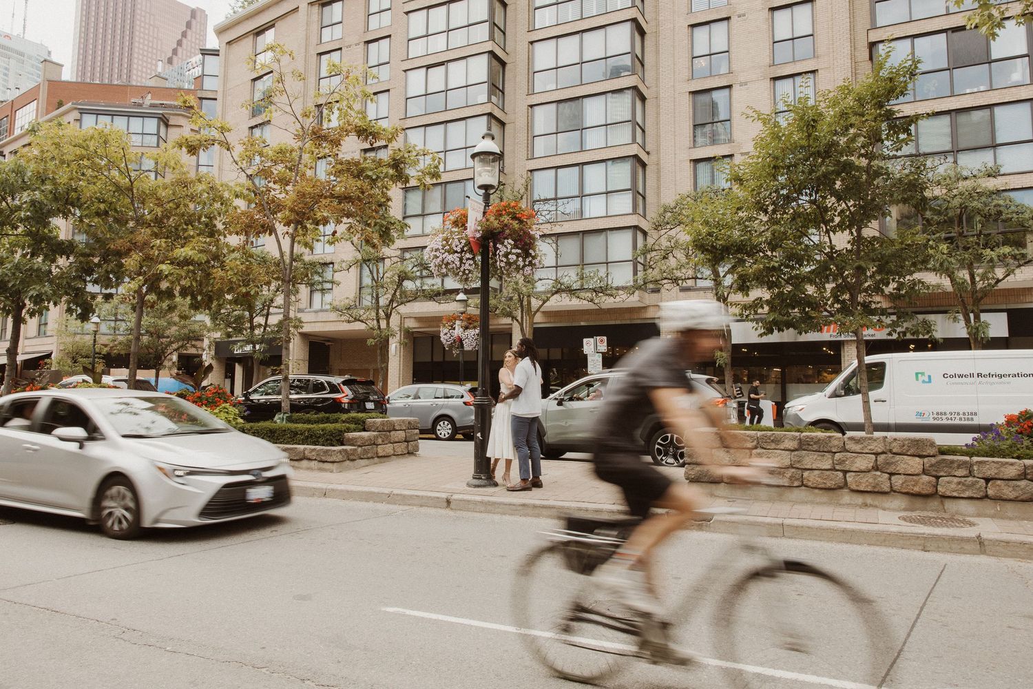 A couple stands near a busy urban street as a cyclist blurs past in front of a luxury hotel.
