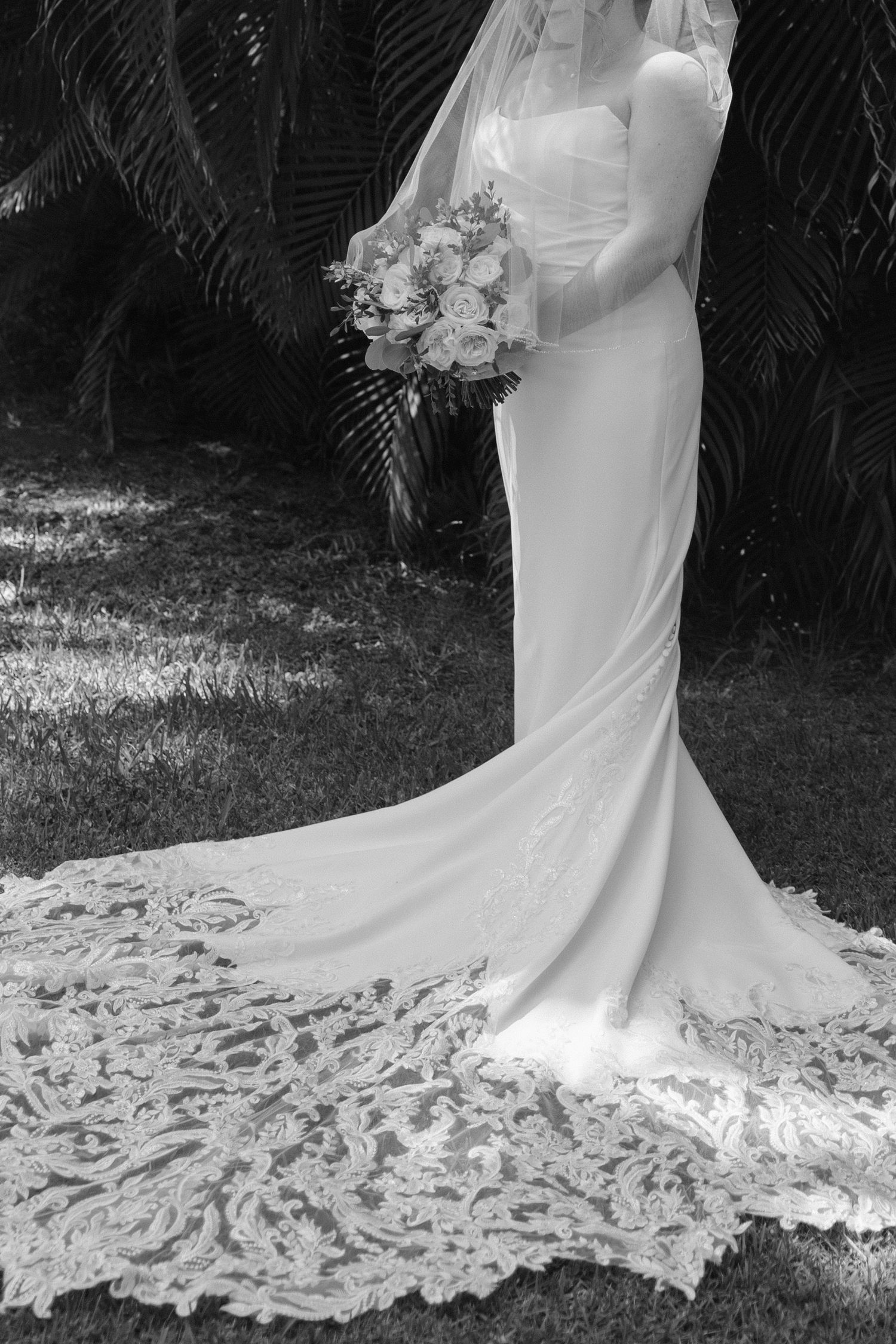 Black and white photo of a bride in a lace-trimmed wedding gown with veil, holding a bouquet outdoors.