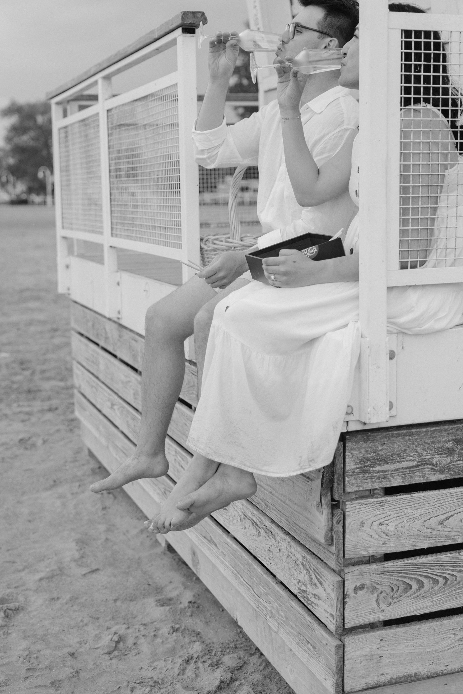 Black and white photo of a woman in white dress sitting on wooden beach hut steps drinking from a bottle.