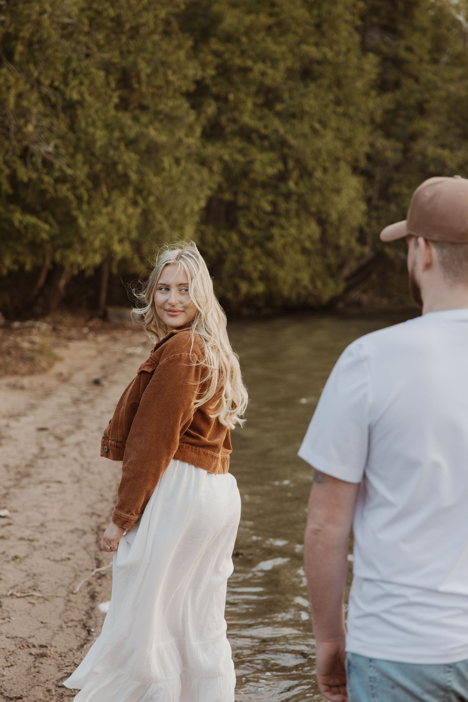 Blonde woman in brown jacket and white skirt smiles over shoulder at man near wooded riverbank.