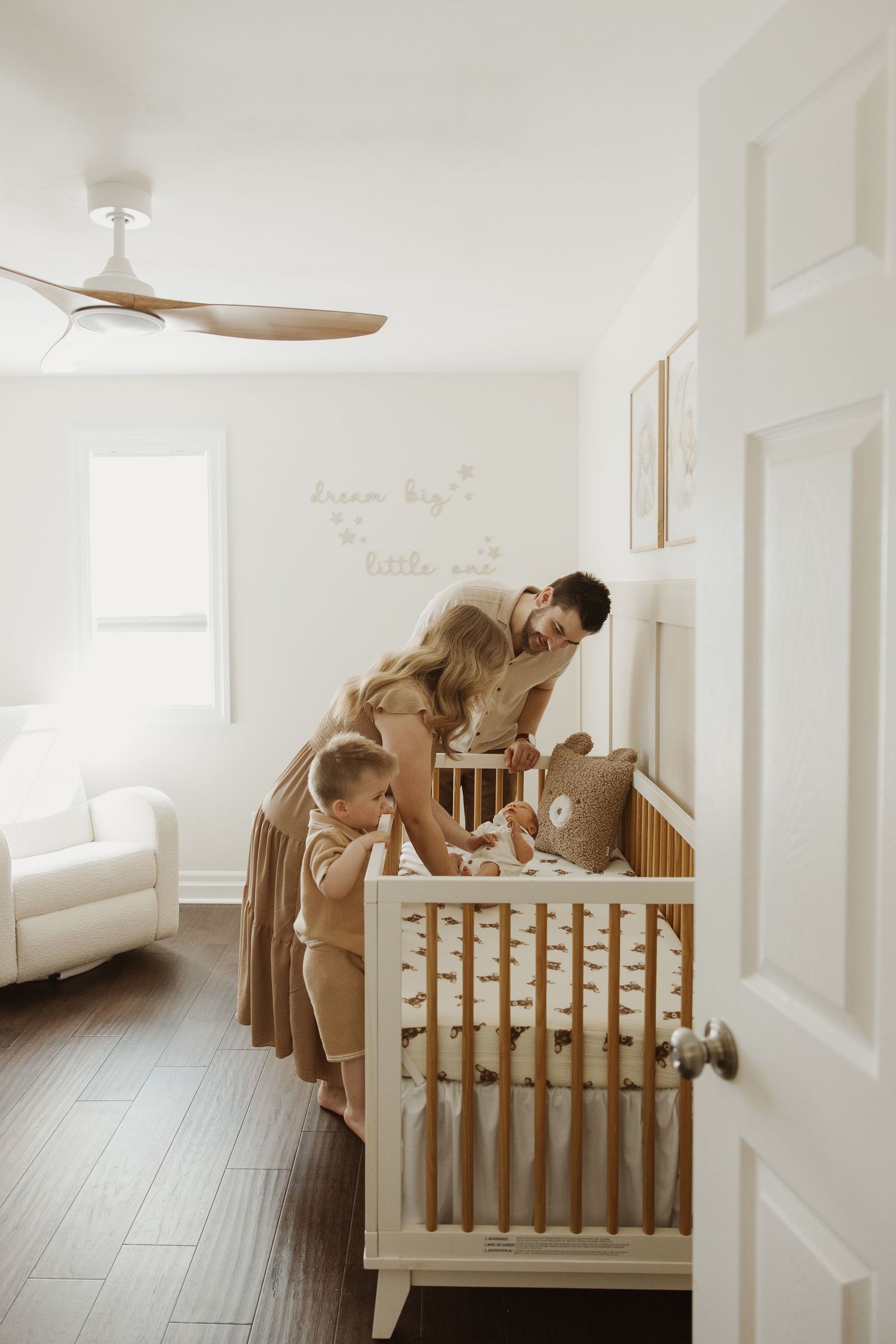 Parents and toddler lean over a wooden crib in a neutral-toned nursery room.