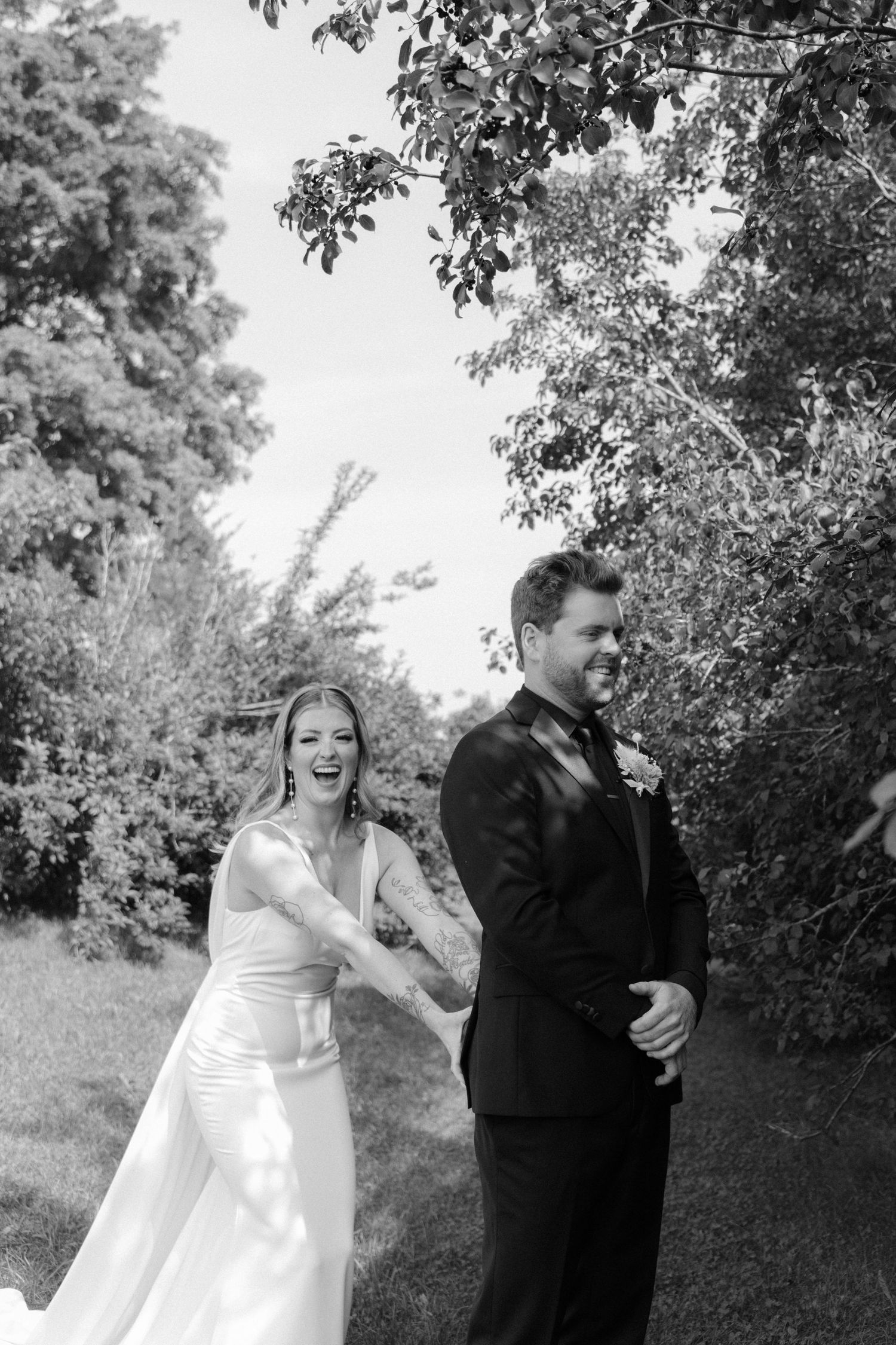 Black and white photo of laughing bride holding groom's arm during a joyful outdoor first look moment.