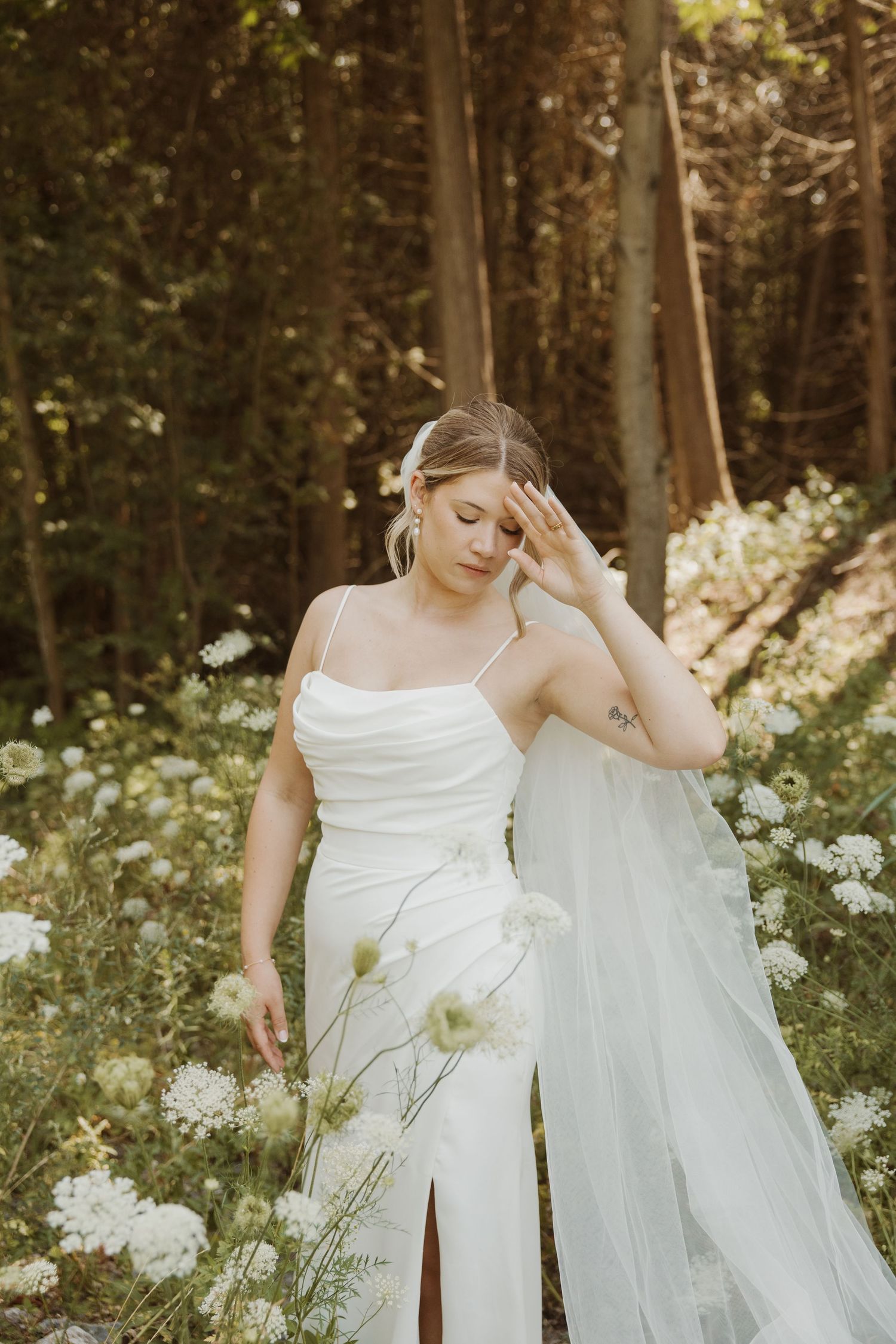 Bride in elegant spaghetti strap wedding dress poses among white wildflowers in sunlit forest.