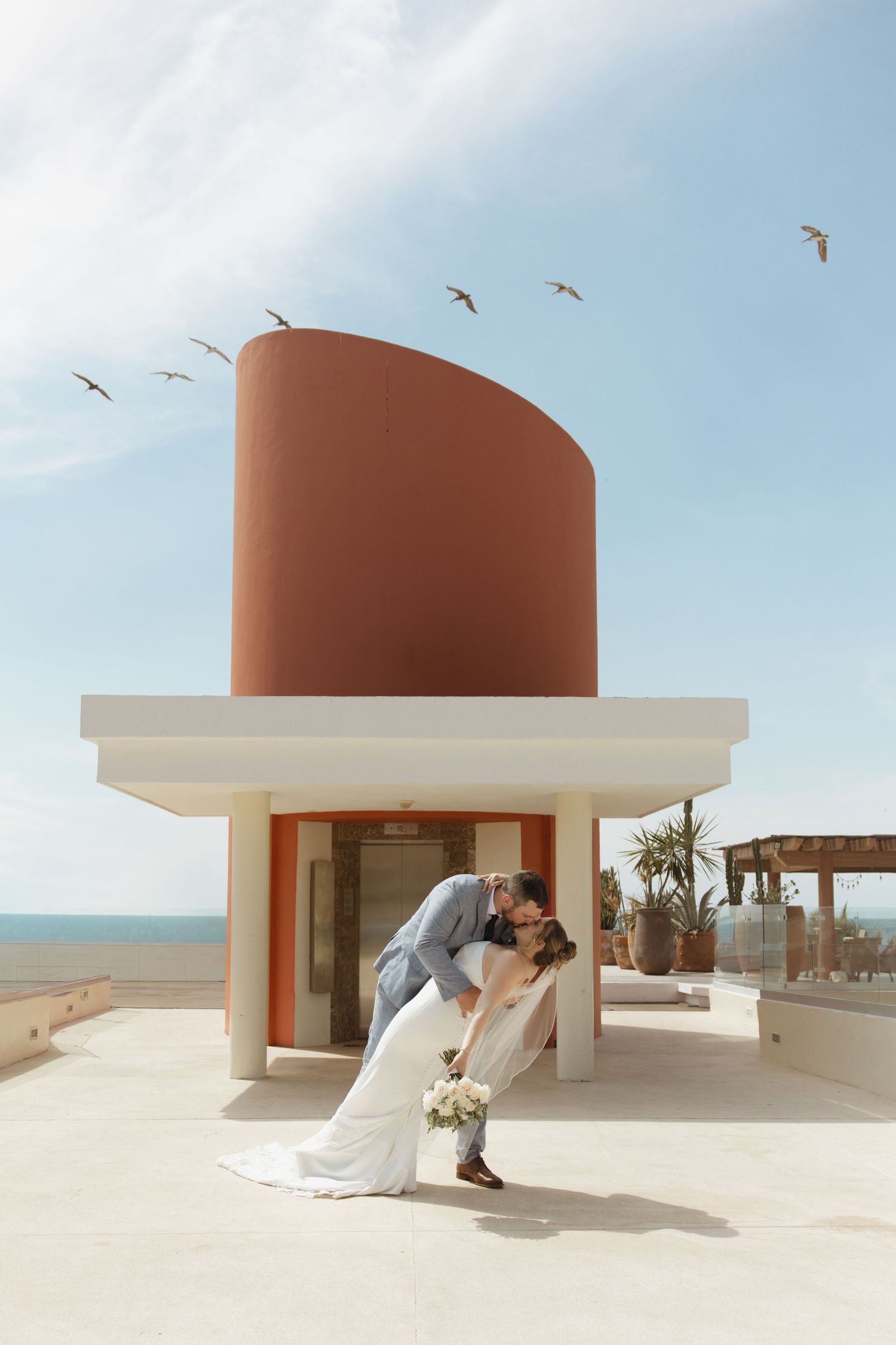 Bride and groom share a romantic dip kiss on a rooftop terrace with birds flying overhead near the ocean.