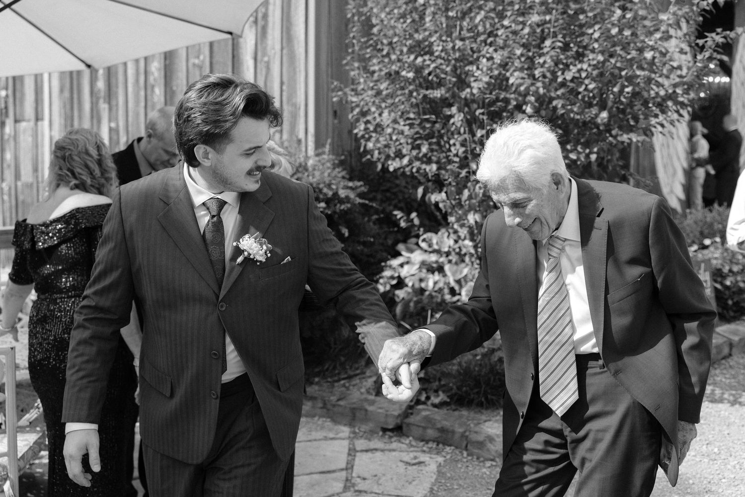 Two men in suits laughing and shaking hands outdoors at a wedding celebration.