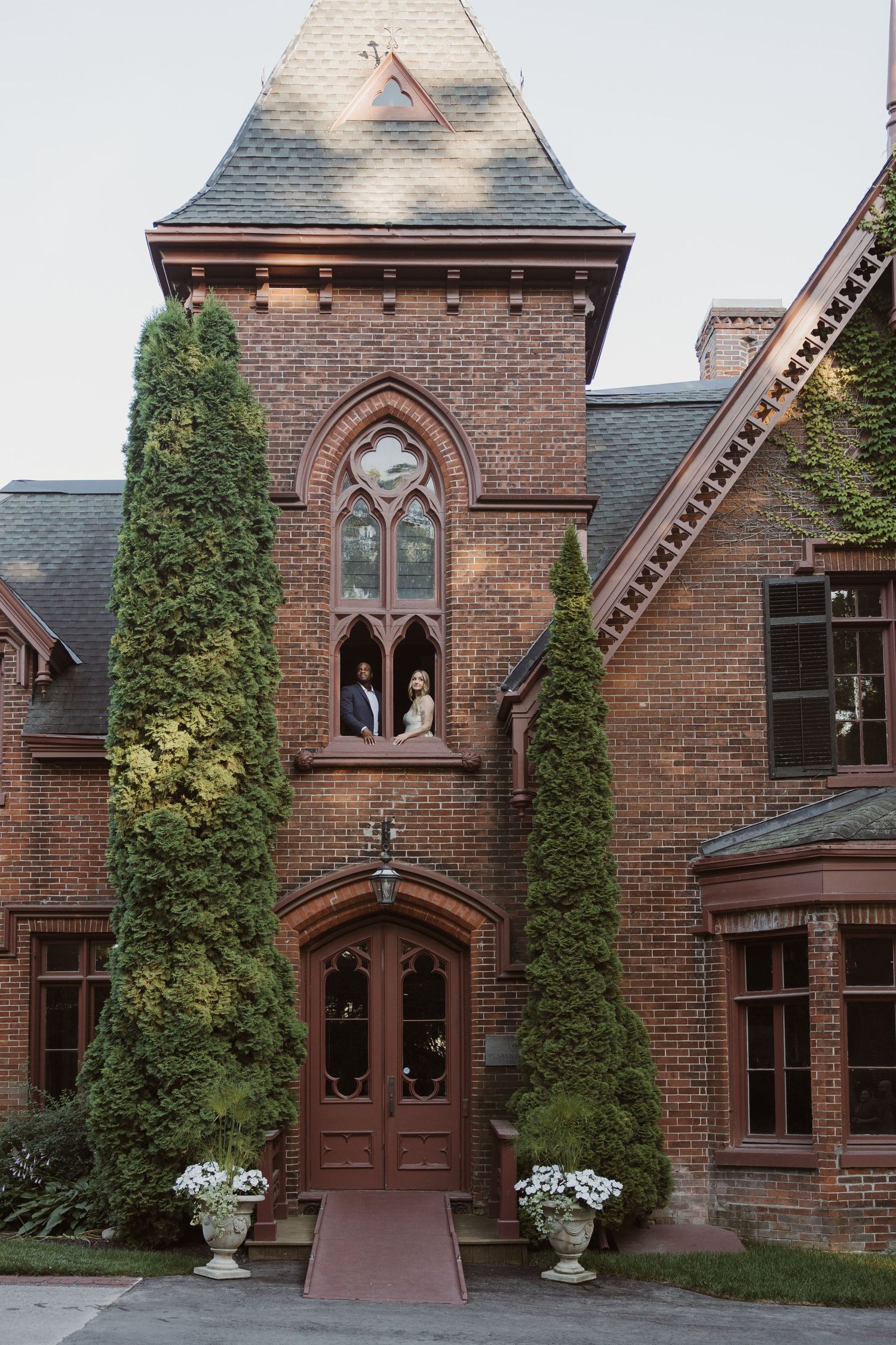 Wedding couple posing in the window of a Gothic brick mansion tower surrounded by tall cypress trees.