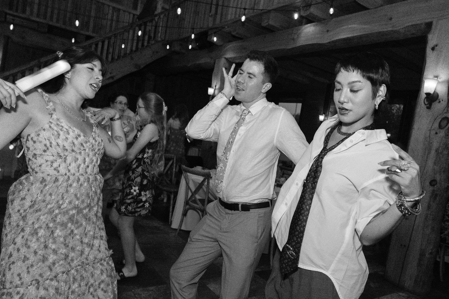 Black and white photo of wedding guests dancing energetically in rustic barn venue with string lights overhead.