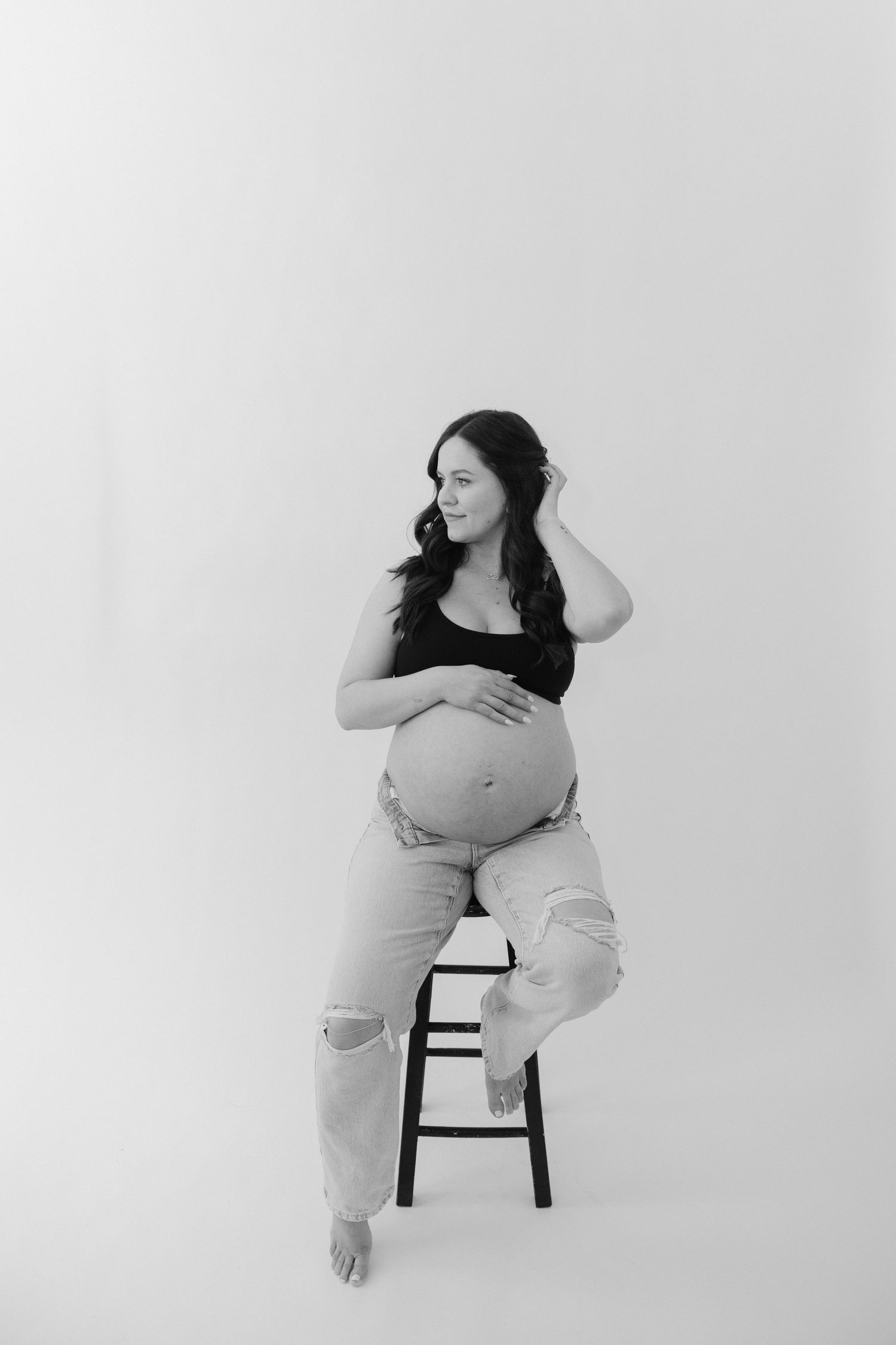 Black and white photo of pregnant woman sitting on stool cradling baby bump.