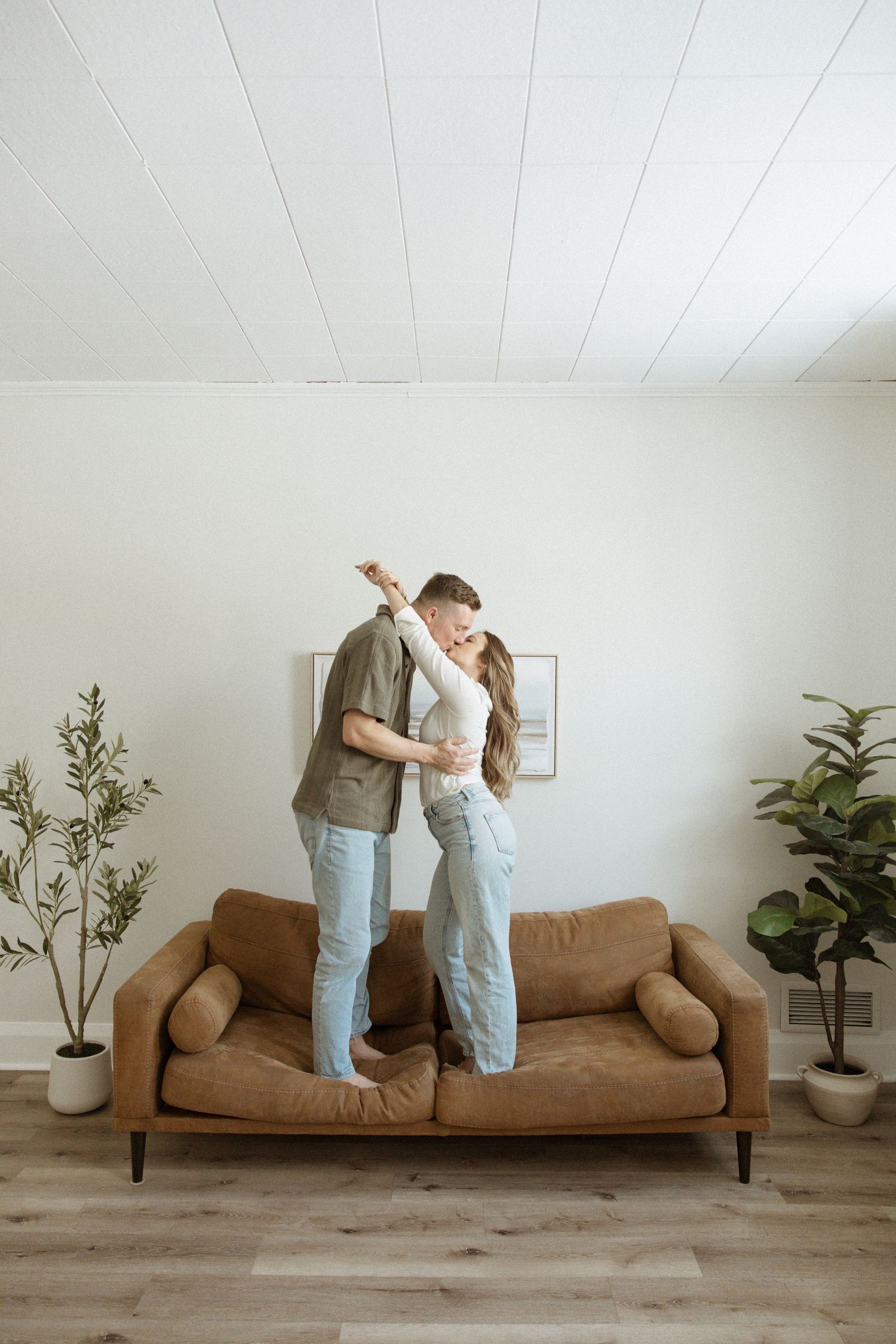 Happy couple sharing a kiss while standing together on a brown leather sofa in a bright living room.