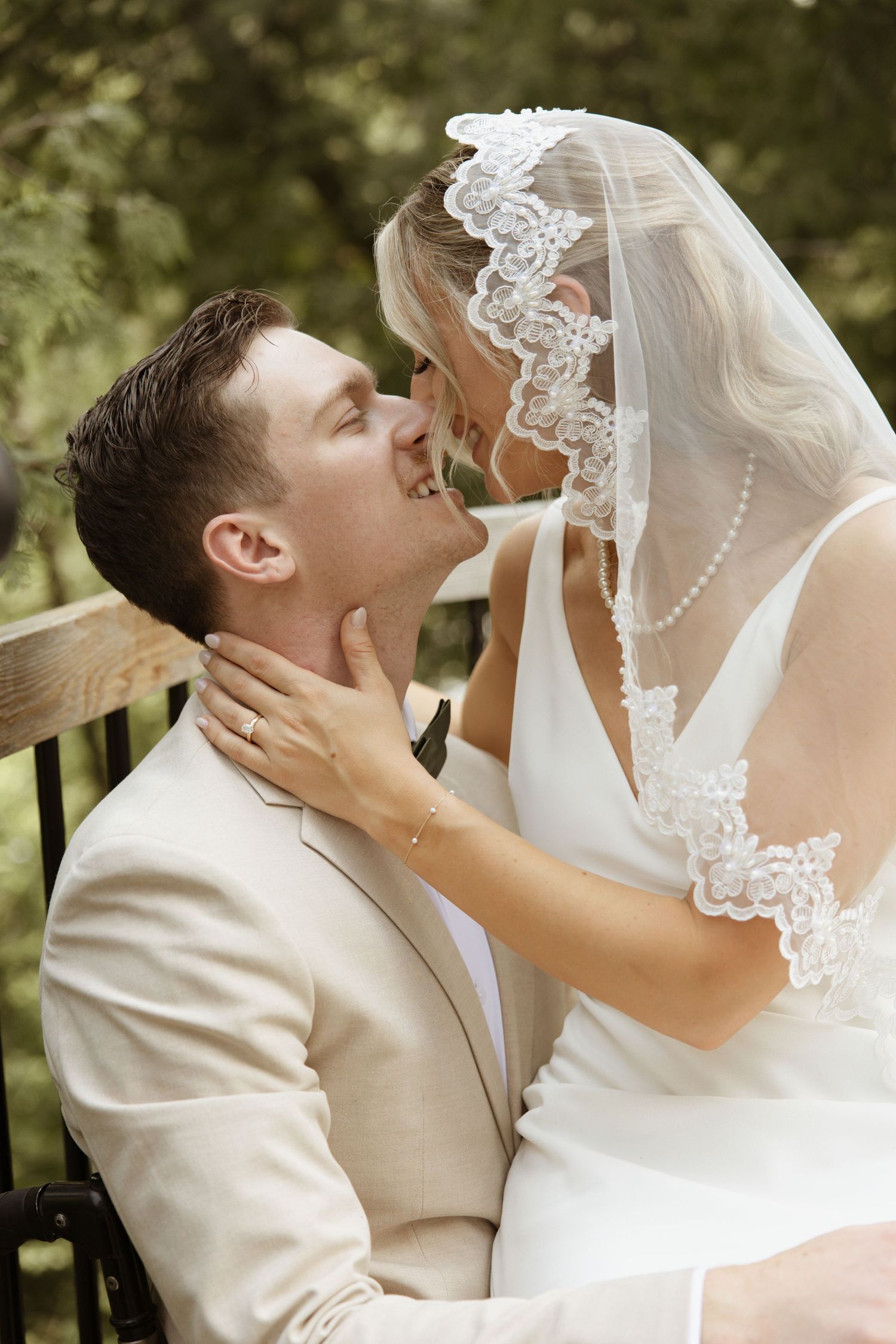 Bride and groom share a romantic almost-kiss on bench, groom in tan suit, bride in lace veil.
