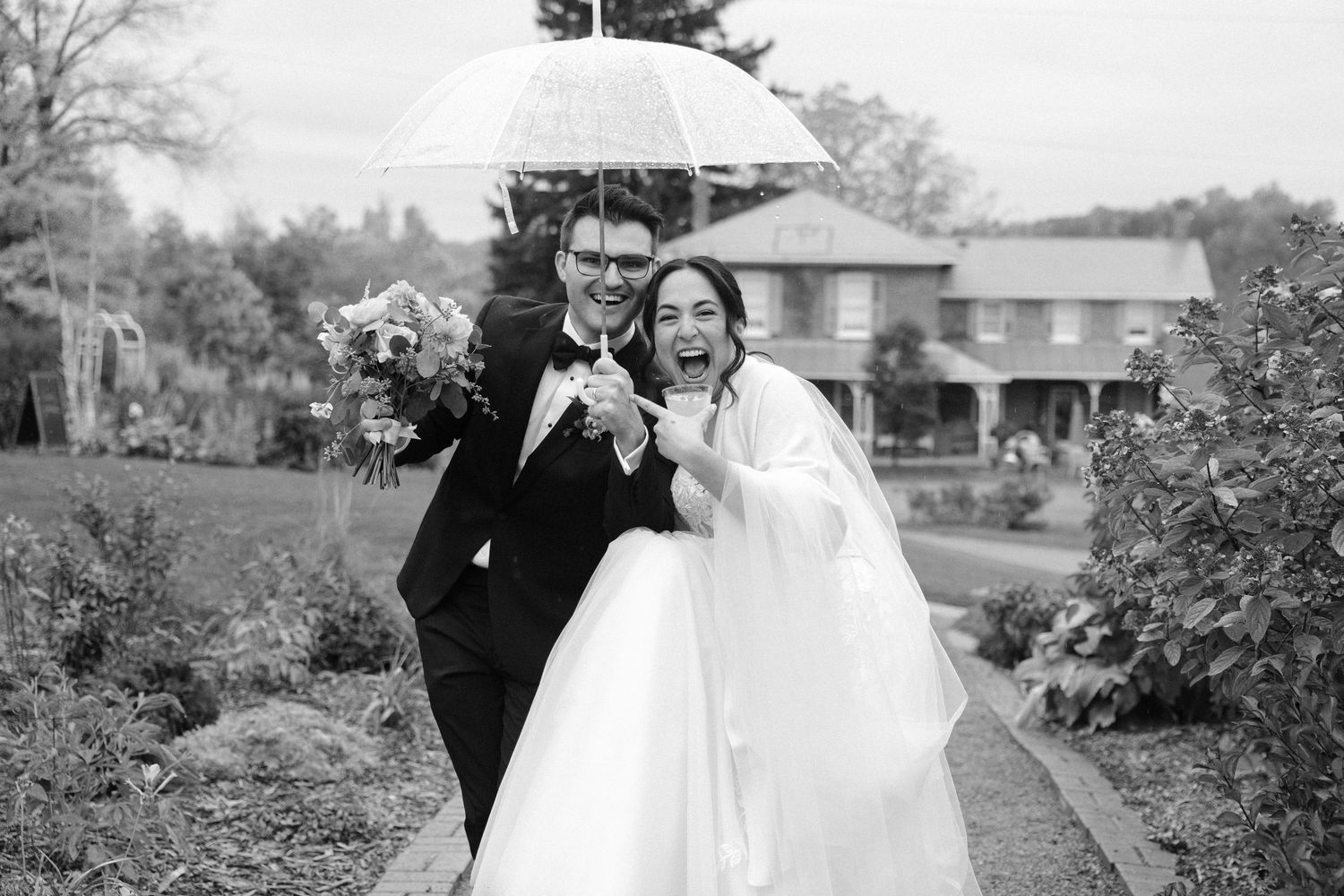 Joyful bride and groom sharing umbrella in garden on rainy wedding day.