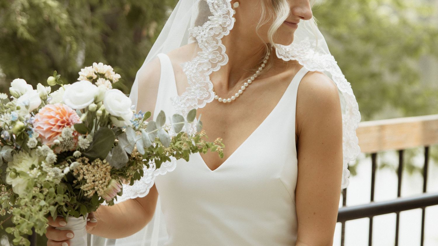 Bride in white gown holding bouquet of roses and greenery wearing lace veil.