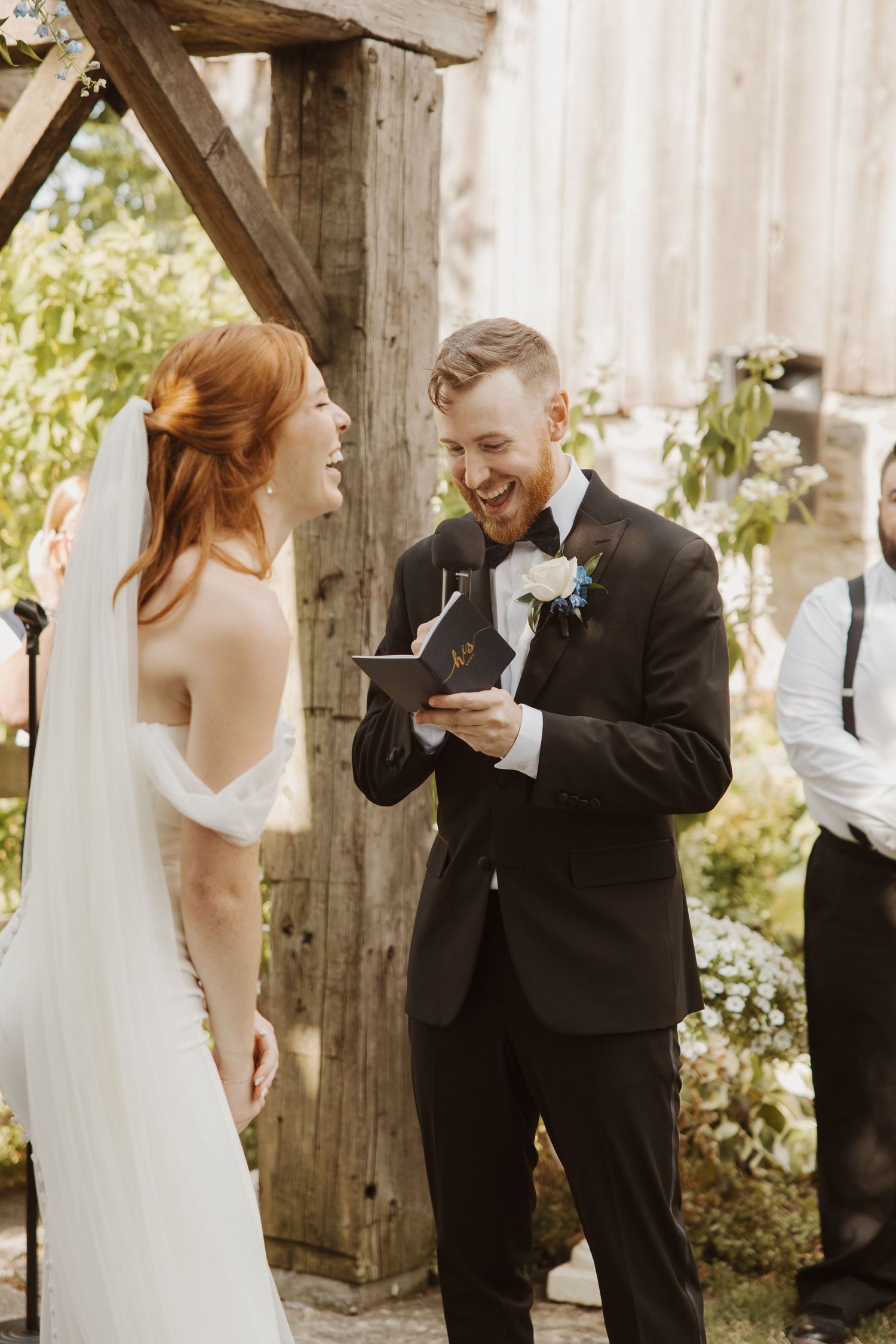 Groom reads vows from book while bride laughs joyfully during outdoor wedding ceremony.